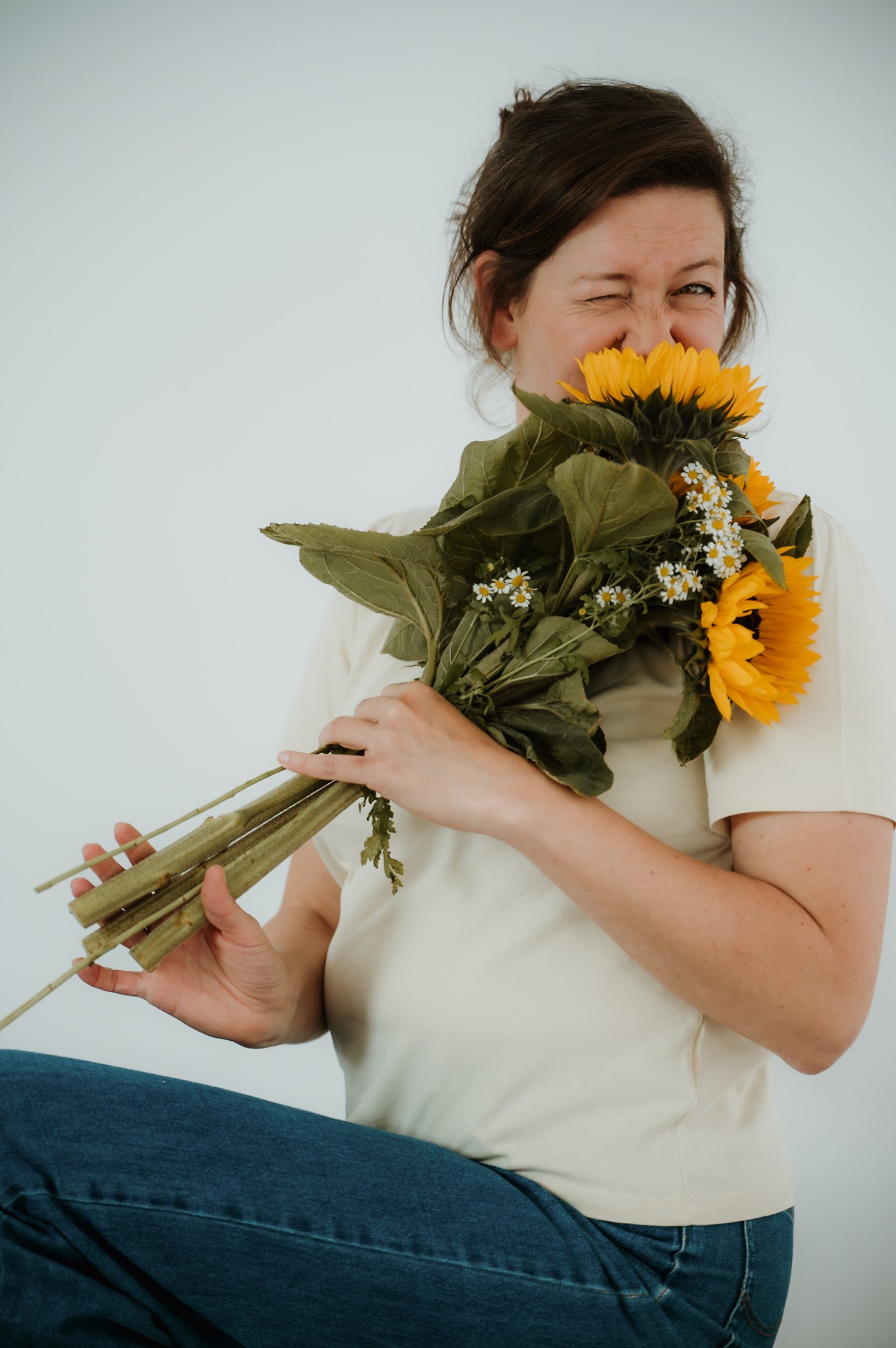 Woman holding a bouquet of sunflowers and small white daisies while smiling and winking, sitting against a plain white background.