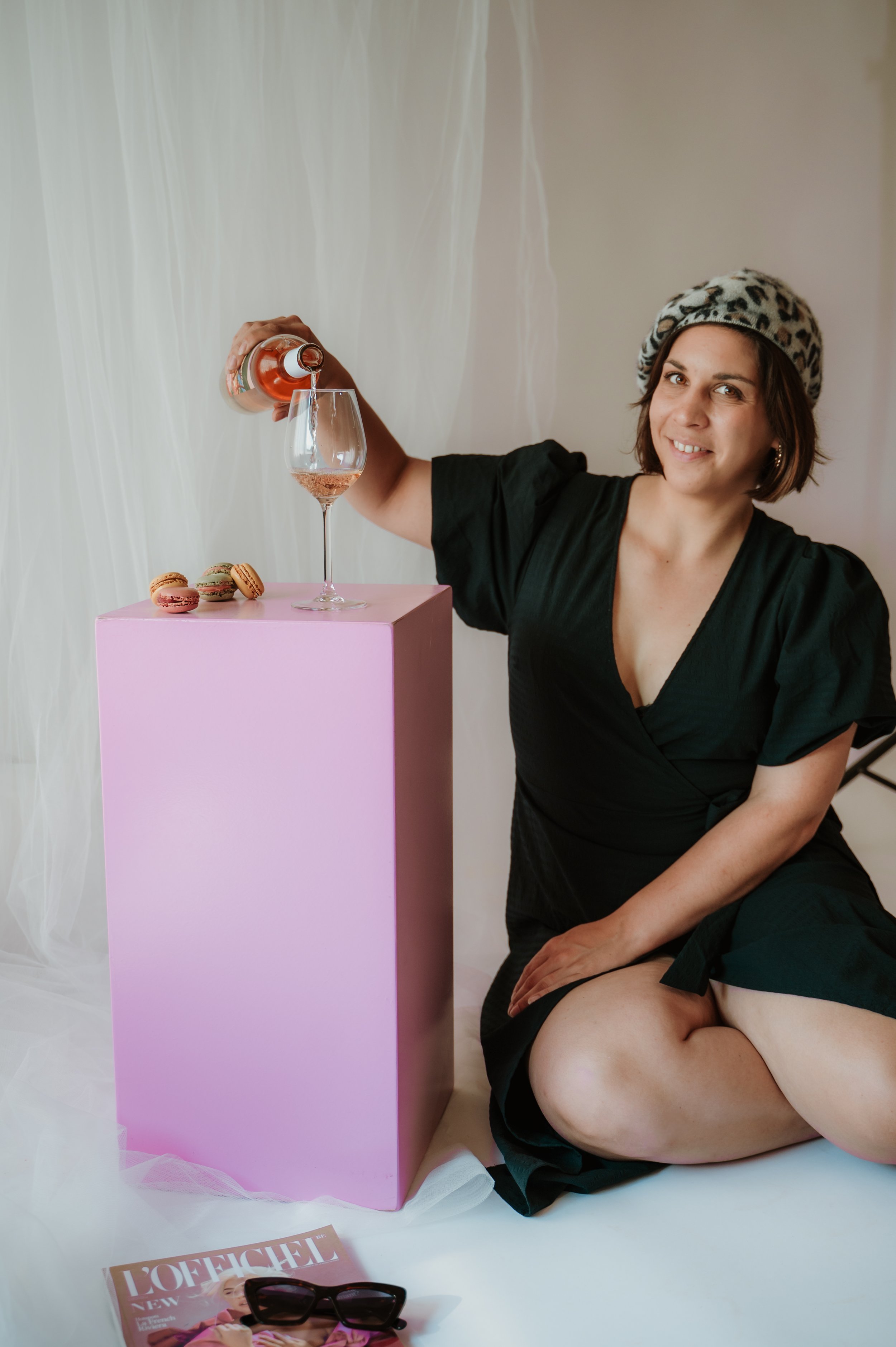 A woman with short brown hair wearing a leopard print beret and black dress, sitting on the floor next to a pink table with a glass of pink-colored beverage and a few macarons, smiling at the camera.
