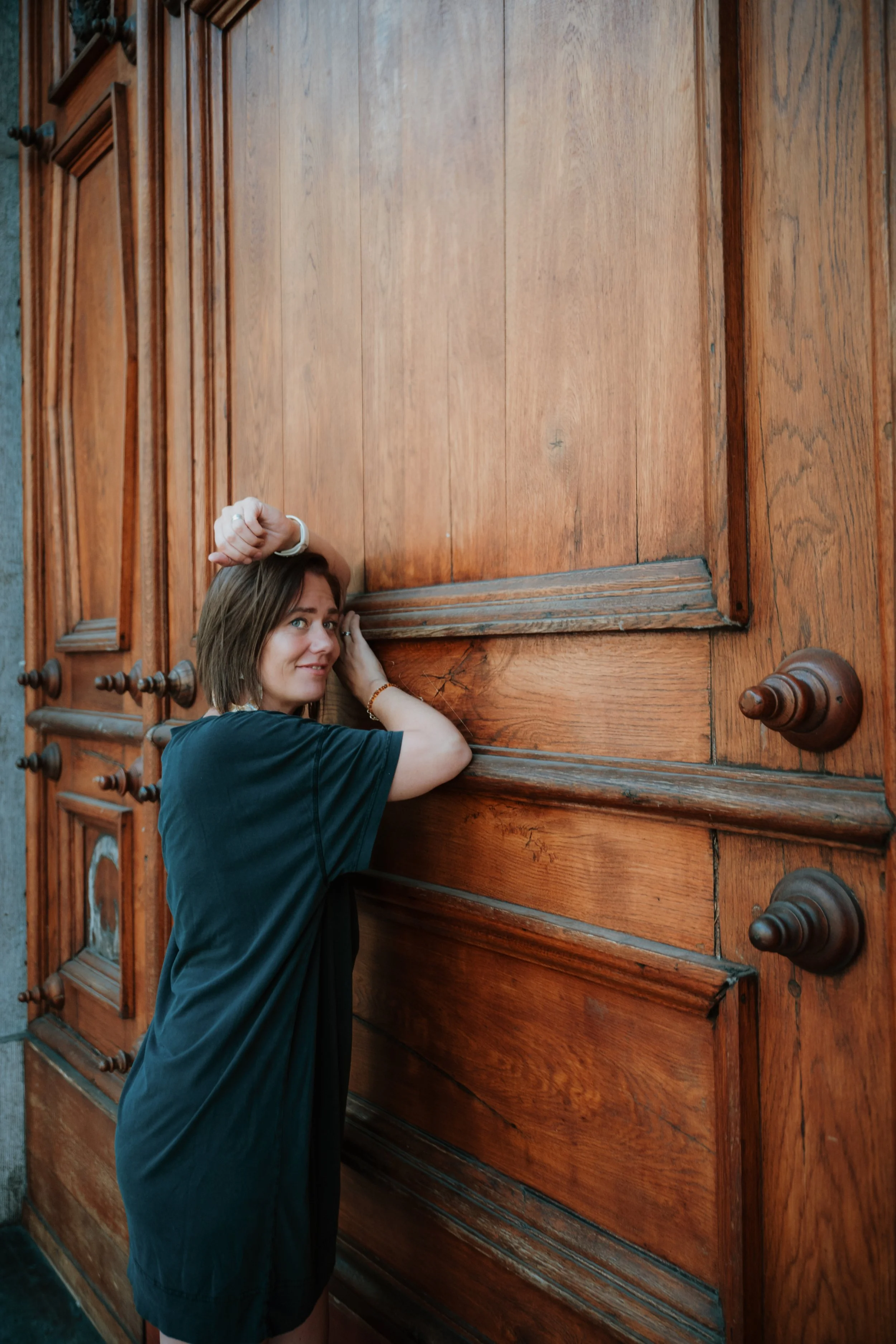 A woman with shoulder-length brown hair, wearing a black t-shirt, is standing beside a large wooden door with intricate carvings. She is resting her arm on the door and looking at the camera with a slight smile.
