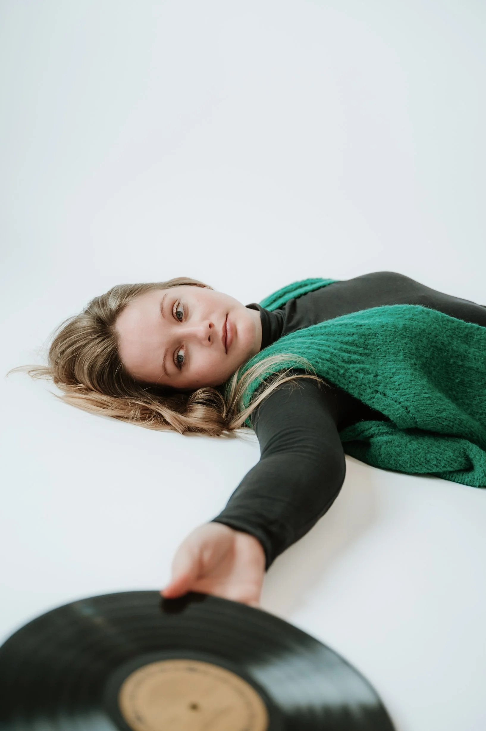 A young woman with long, wavy blonde hair lying on the floor, reaching towards the camera with her right arm. She is wearing a black top and a green knitted vest, situated against a plain white background.