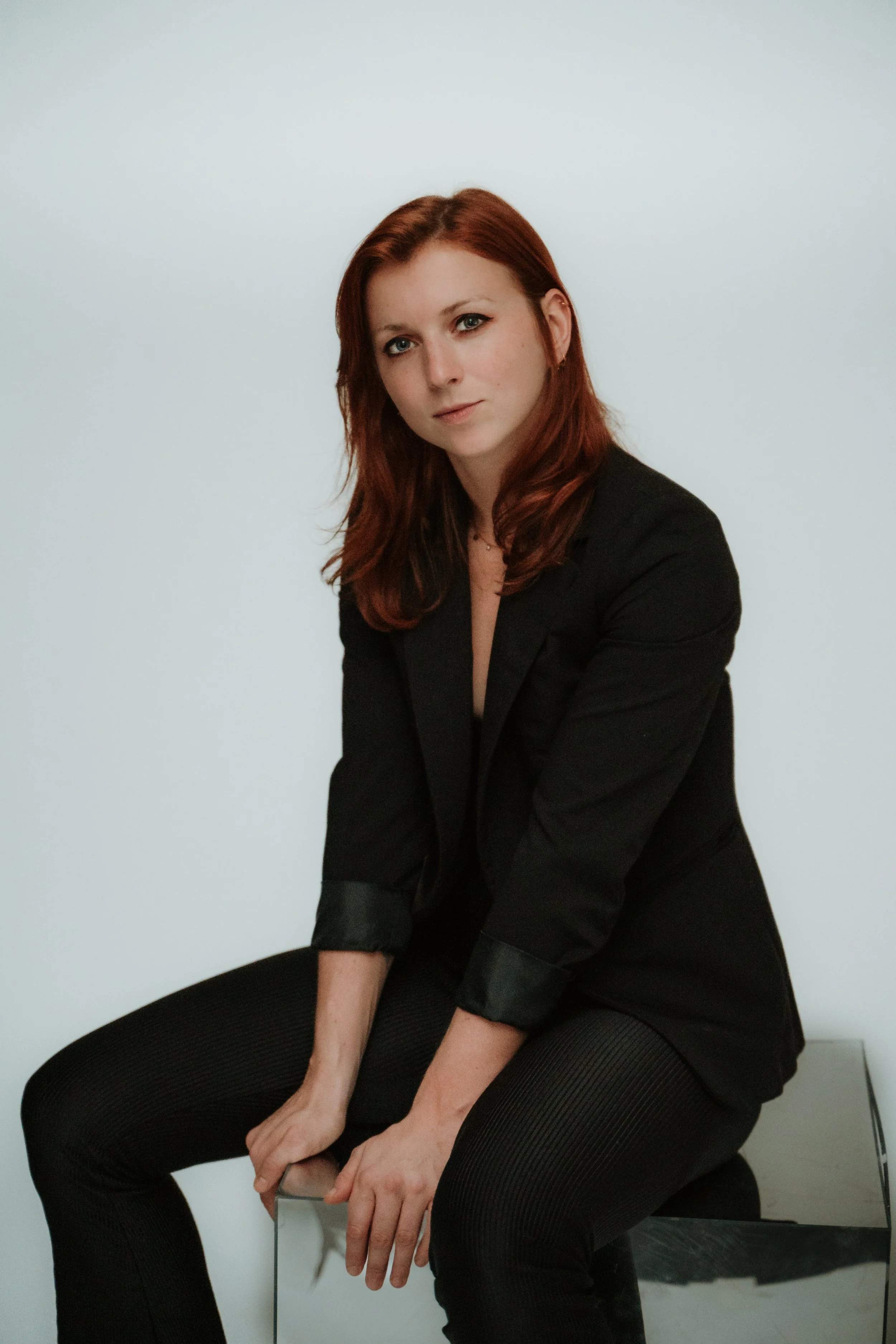 A woman with red hair sitting with her hands on her knees, wearing a black jacket and black pants against a plain white wall.