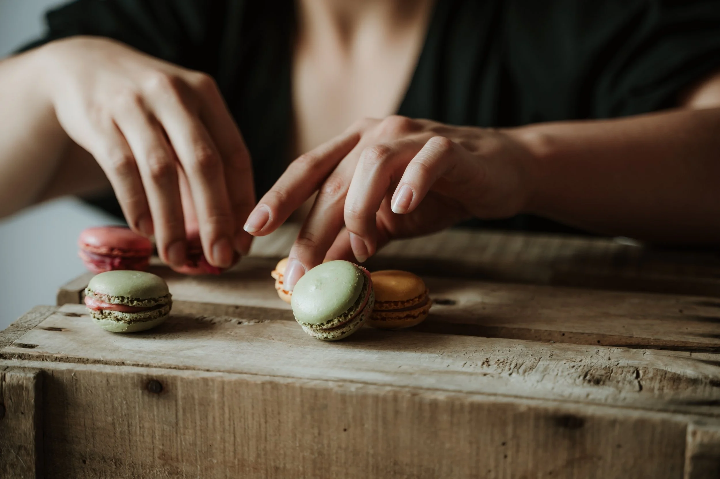 Close-up of a person’s hands arranging colorful macarons on a wooden surface.