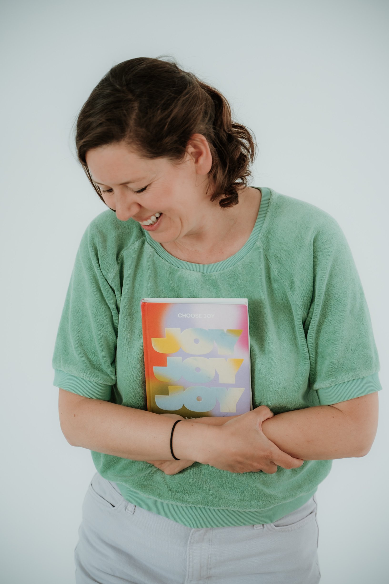 A woman with short brown hair wearing a light green shirt and white pants holding a colorful booklet that reads 'Joy' multiple times, standing against a plain light gray background.