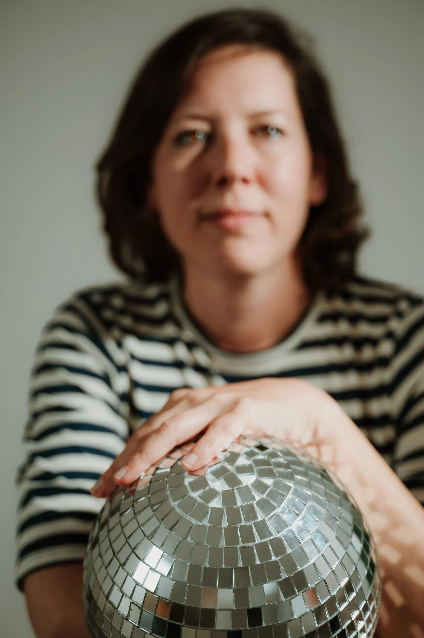 A woman with shoulder-length brown hair wearing a black and white striped shirt, holding a disco ball covered with small mirror tiles.