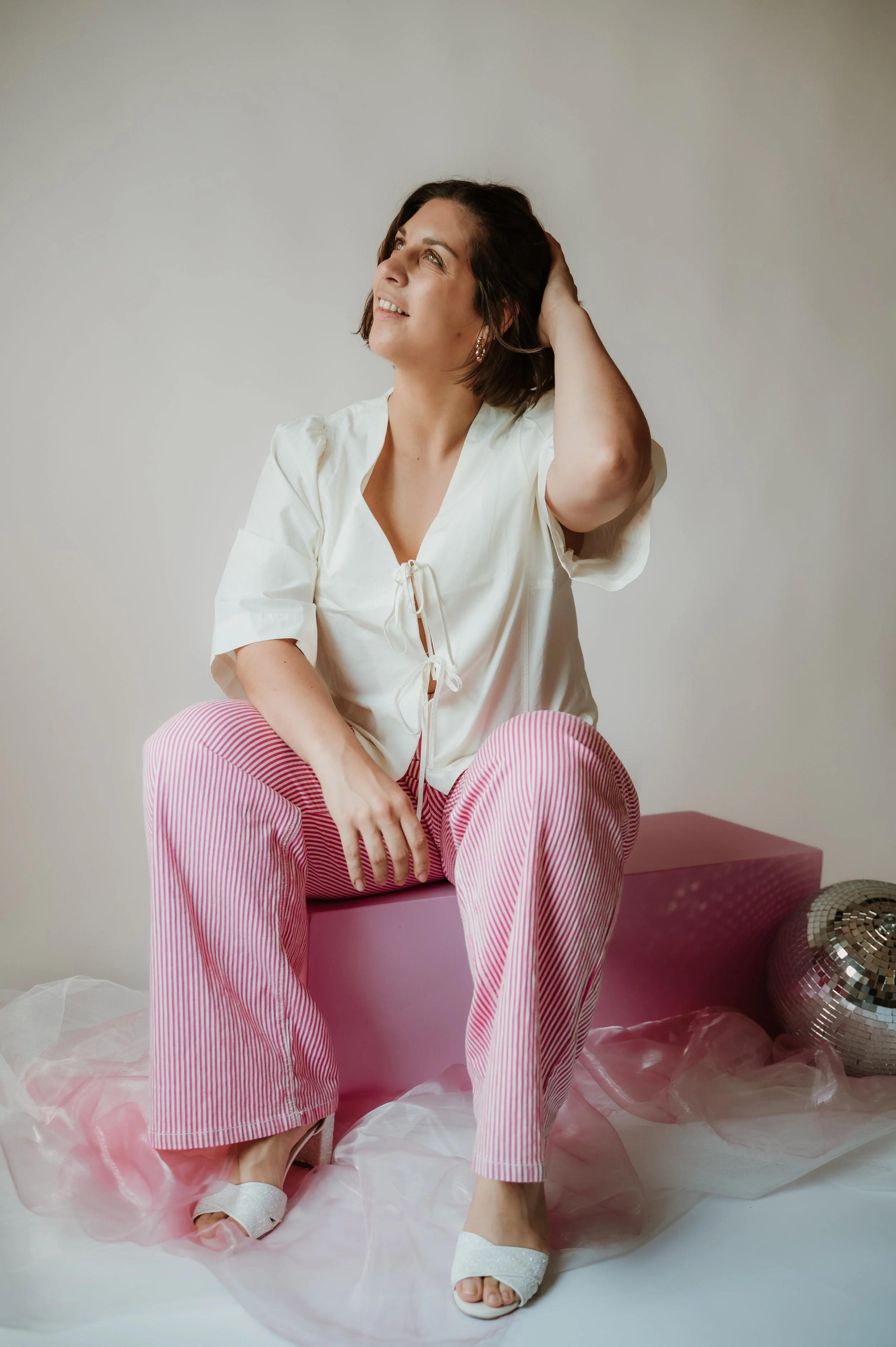 A woman sitting on a pink block, wearing pink and white striped pants, a white top, and white heels, surrounded by pink and white tissue paper and a large silver balloon.