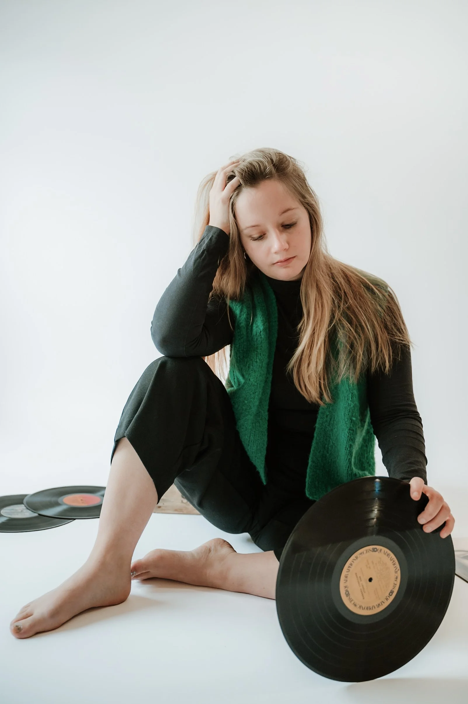 A young woman with blonde hair sitting on the floor, holding a vinyl record, with another record on the floor beside her, against a white background.