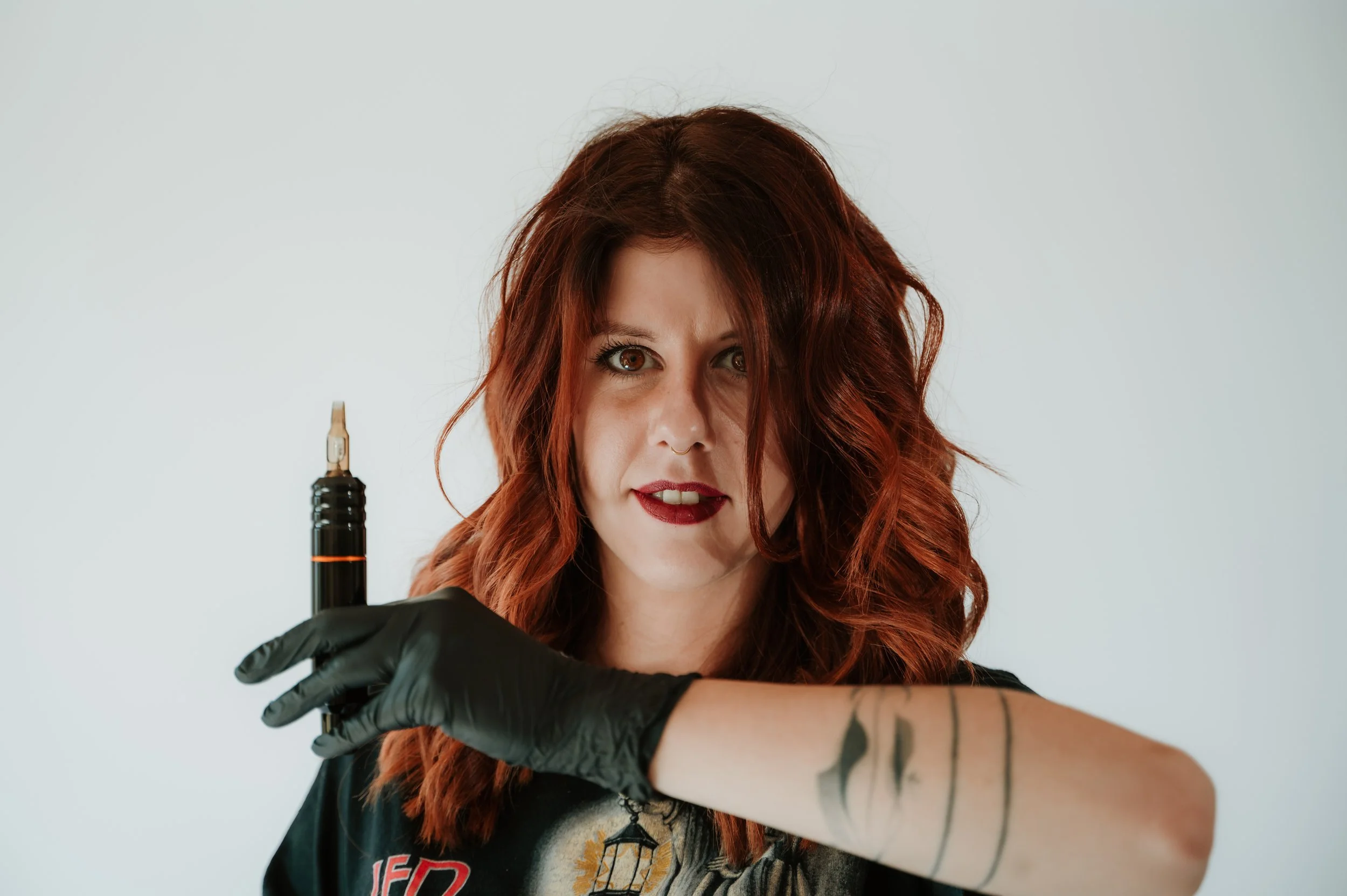 A woman with red hair and tattoos on her arm, wearing black gloves, holding a tattoo machine, and looking into the camera.