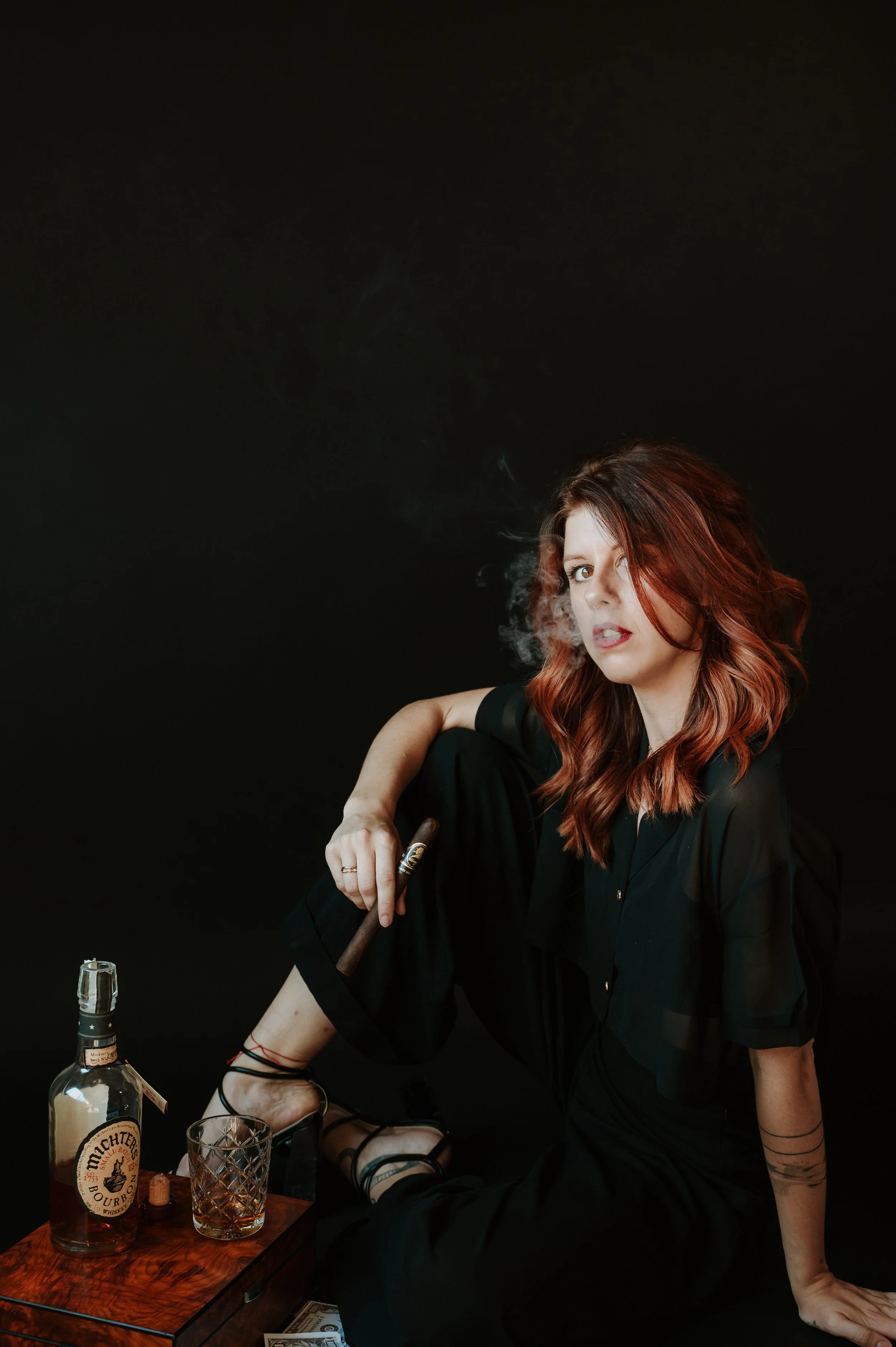 Woman with red hair and tattoos sitting on a table, smoking a cigar, with a bottle of alcohol and a glass in front of her, against a black background.