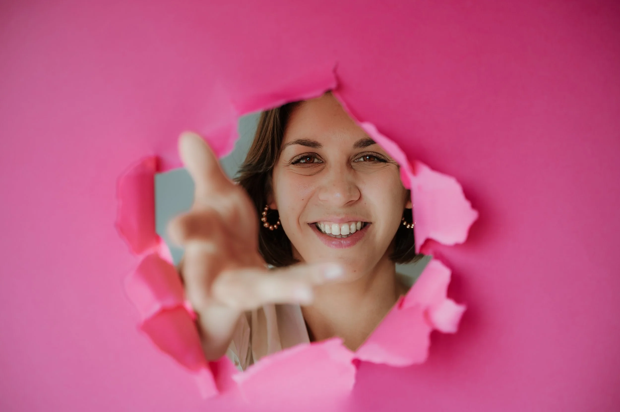 Woman smiling gives a thumbs-up through a torn pink paper.