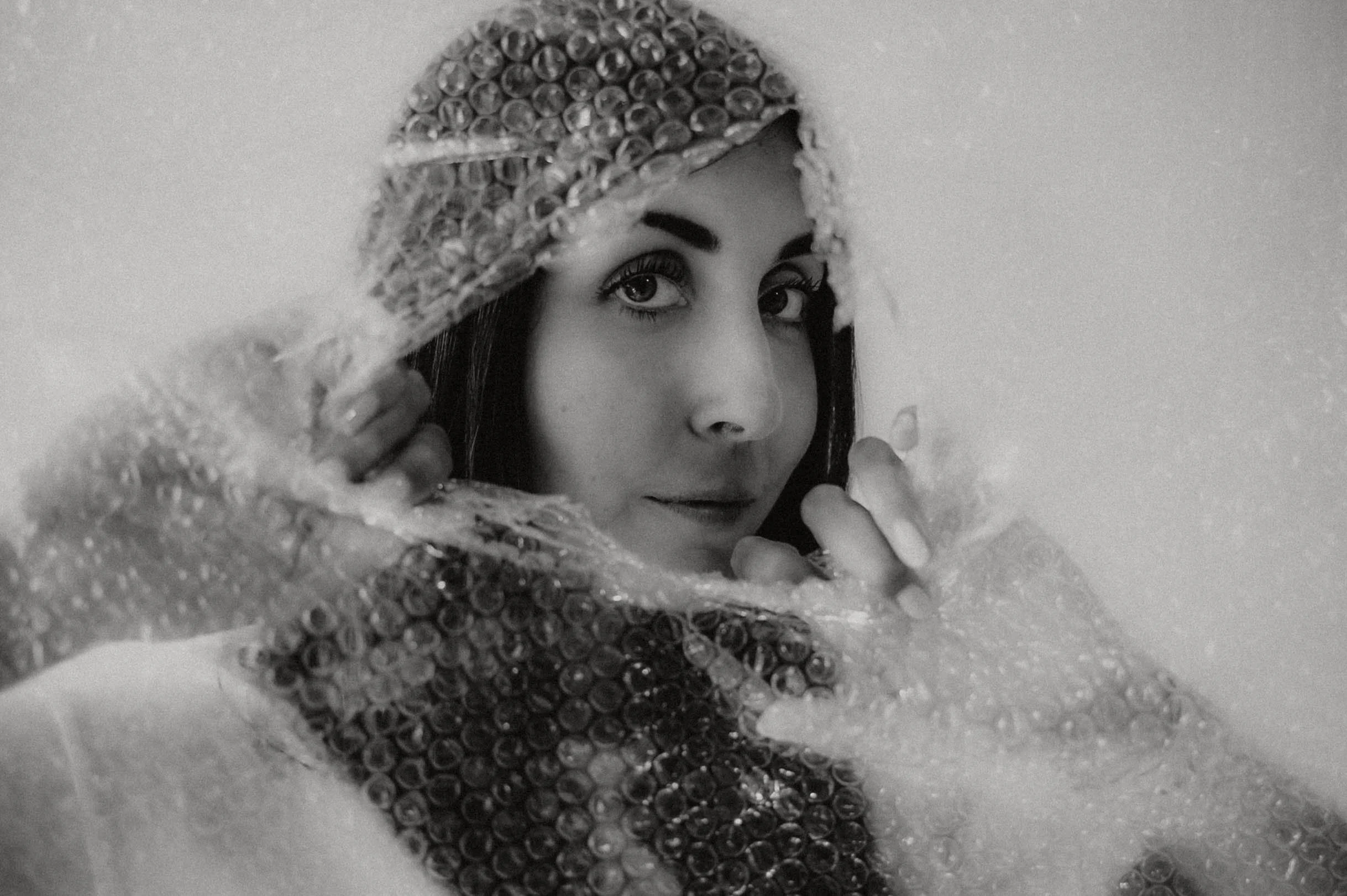 A woman with dark hair and dramatic eye makeup looks directly at the camera through a circle of bubbles or beads, partly covering her face.