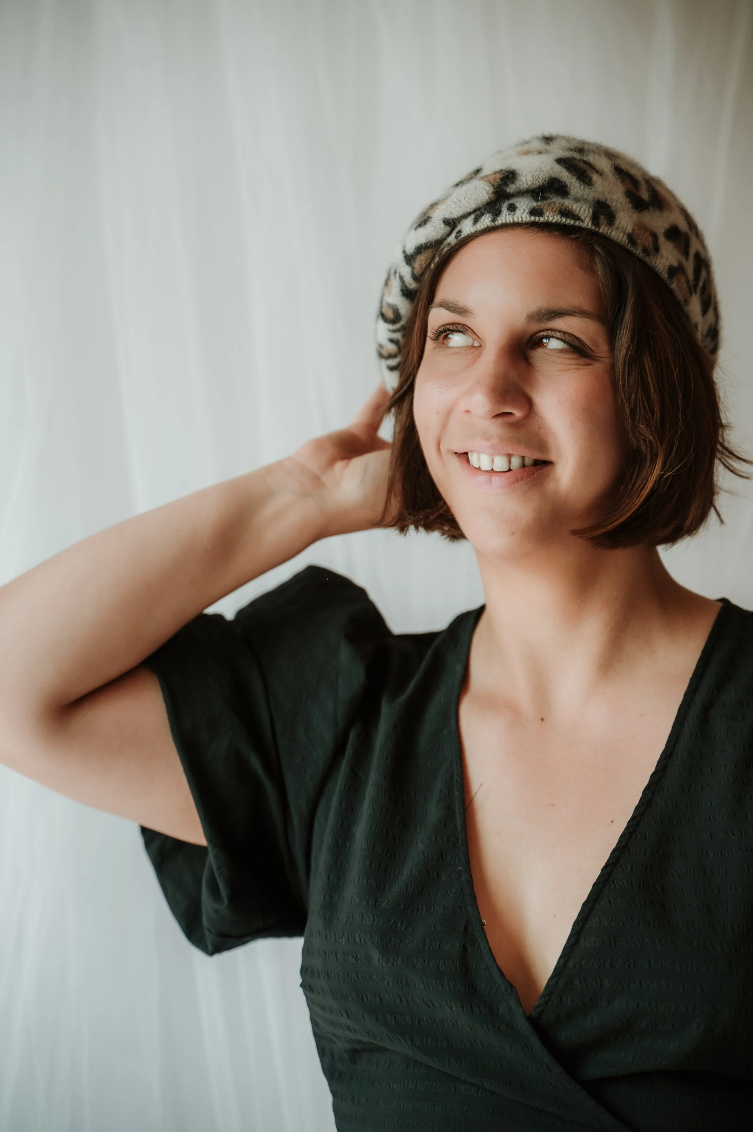 A young woman with short brown hair wearing a leopard print beret and a black textured dress, smiling and looking to the side against a plain white background.