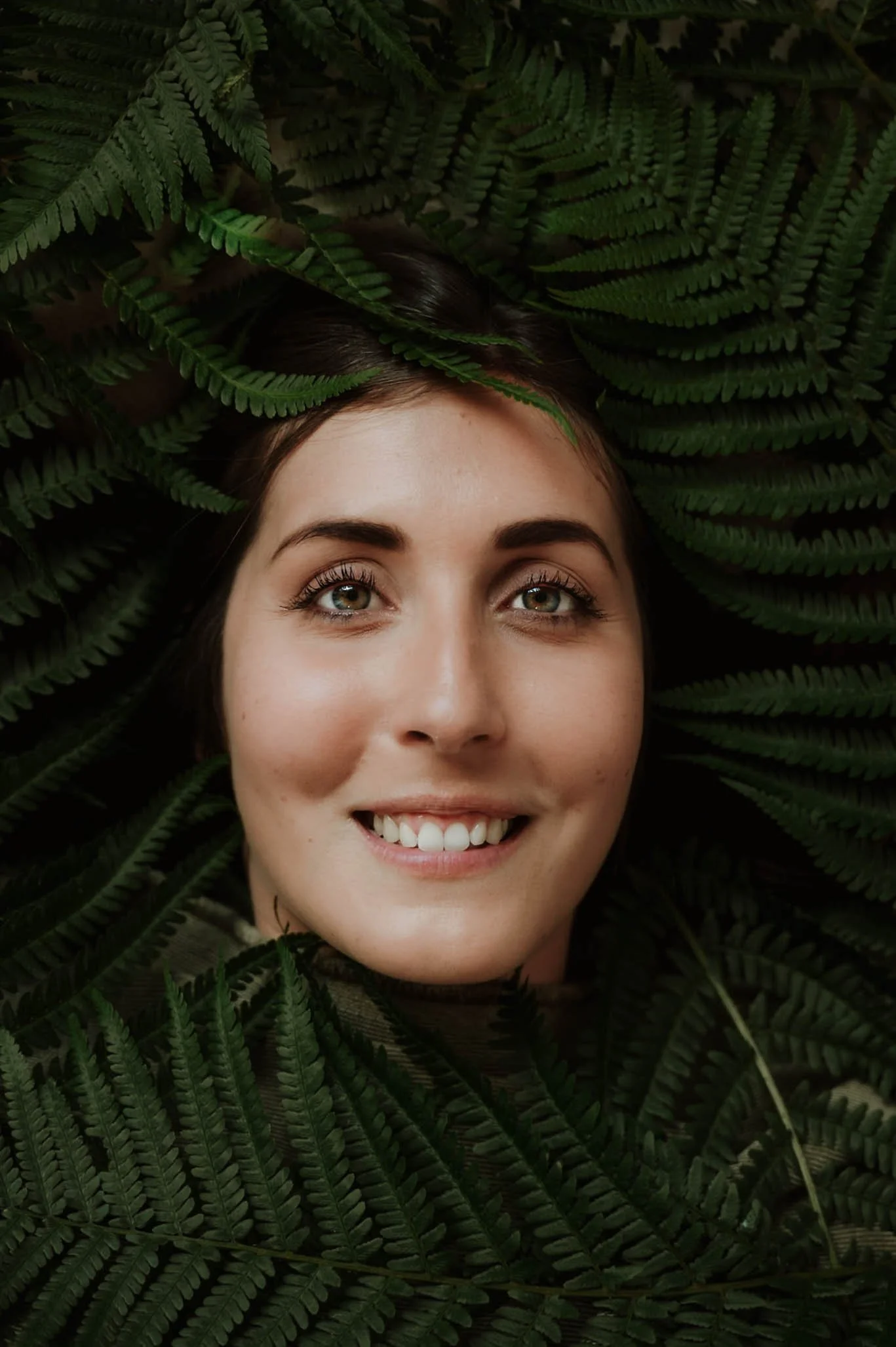 Close-up of a smiling woman with light skin and brown hair, surrounded by lush green fern leaves.