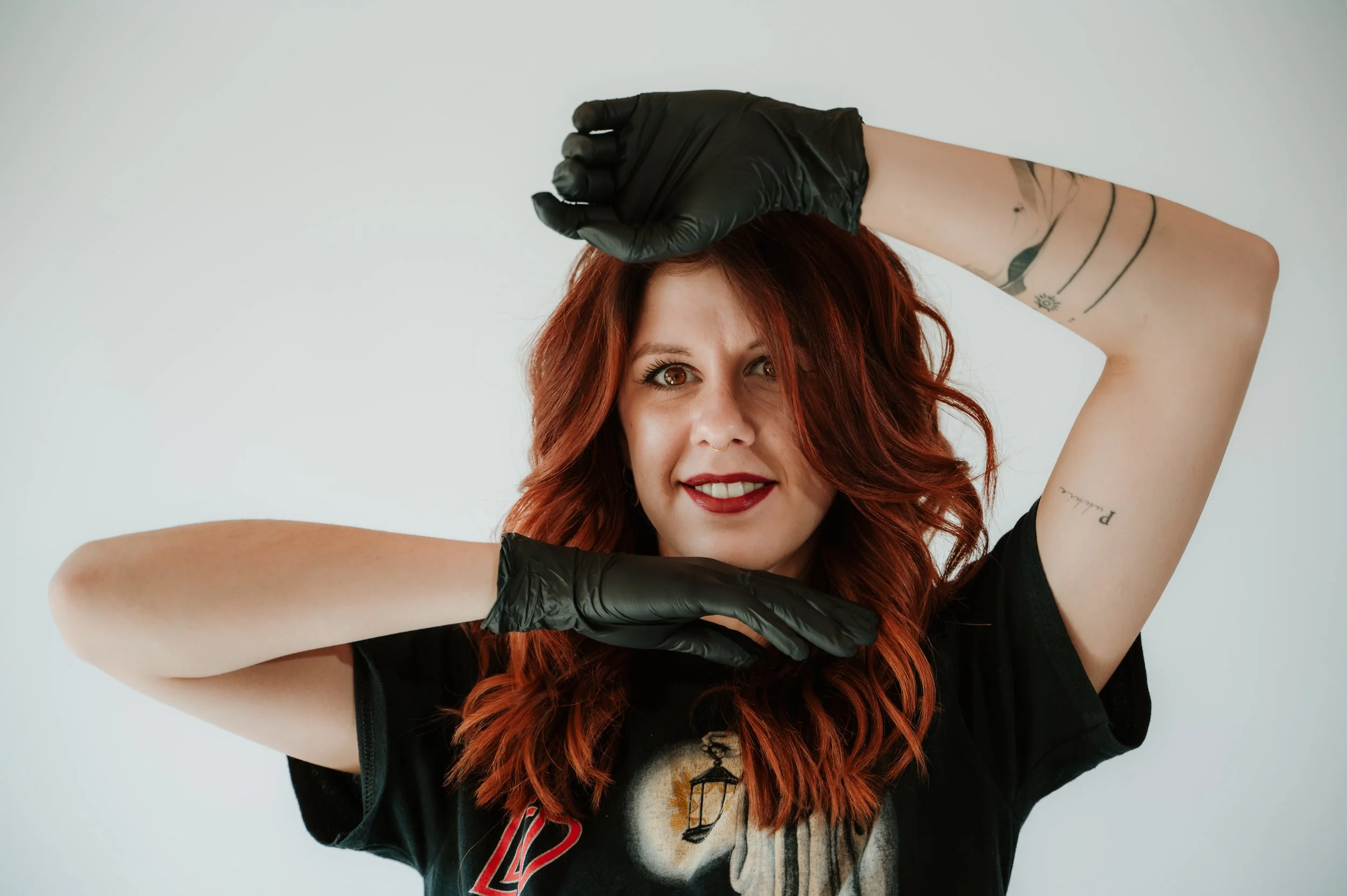 Young woman with red hair, smiling, wearing a black glove on her right hand, and a black T-shirt with a graphic design, against a plain light gray background.