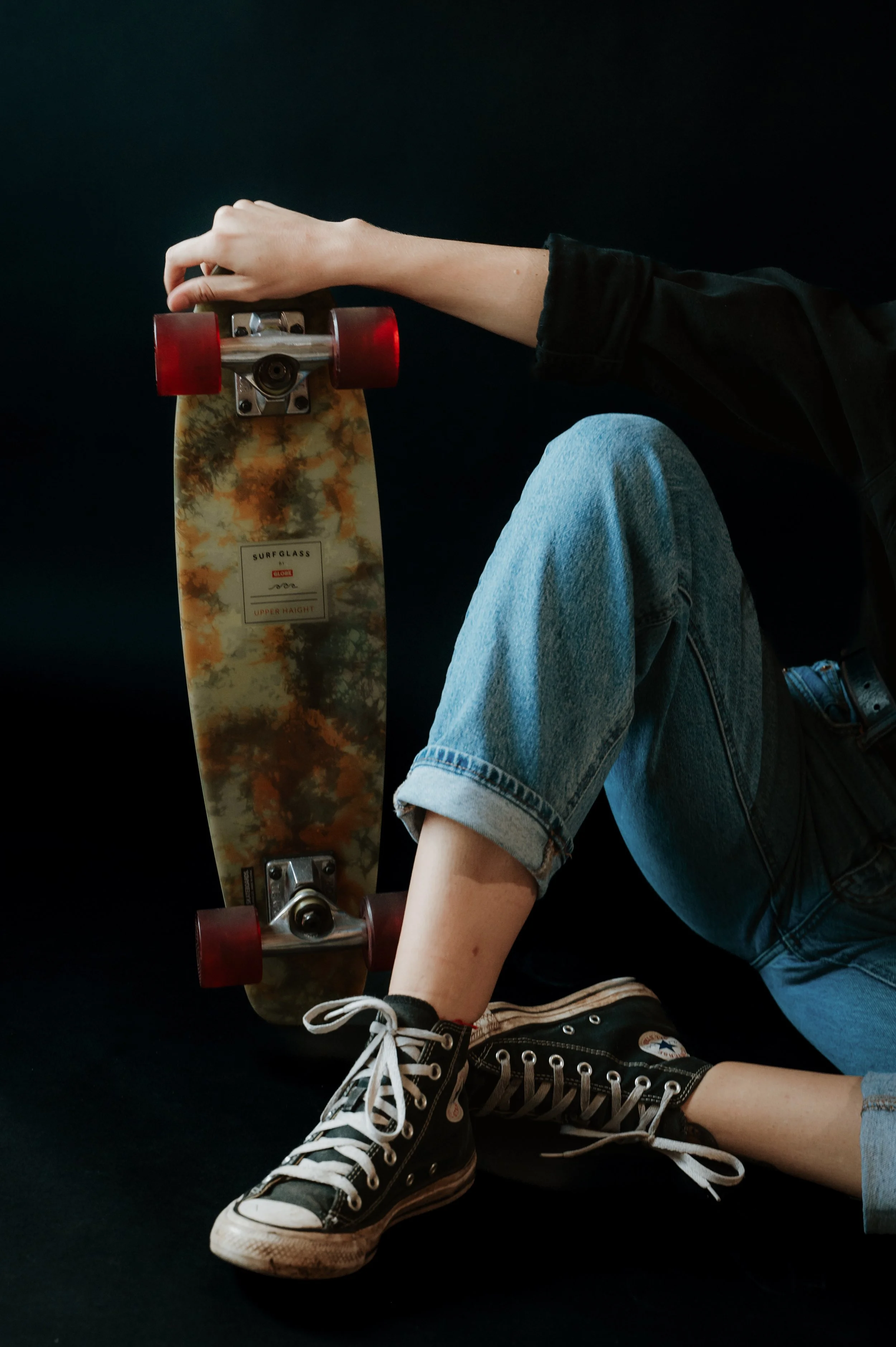 Person sitting against a black background, wearing blue jeans and black Converse sneakers, with their left arm holding a skateboard upright.