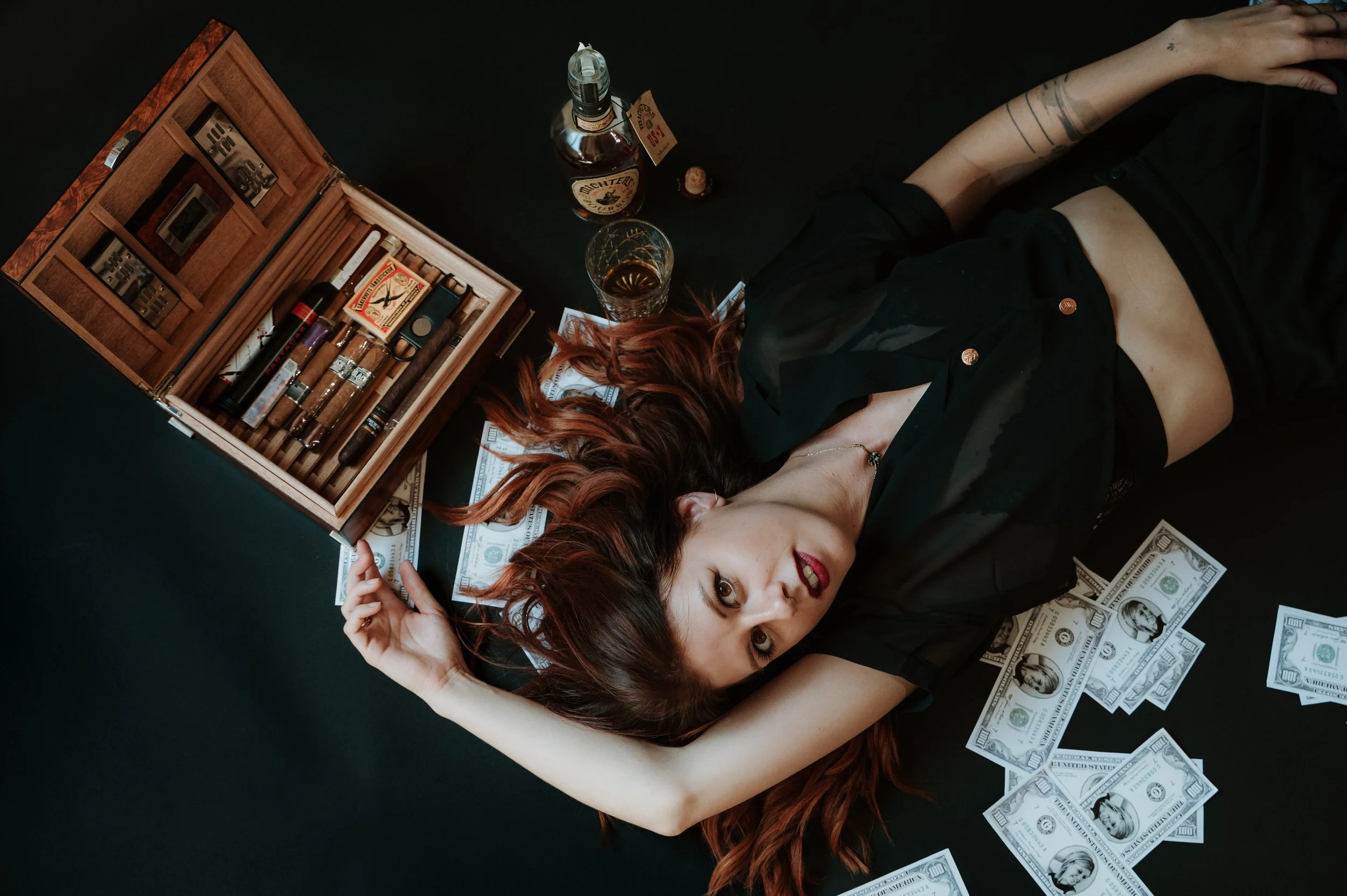 A woman with red hair lies on a black surface surrounded by cash, a wooden cigar humidor with cigars, a glass of alcohol, and a bottle of liquor.