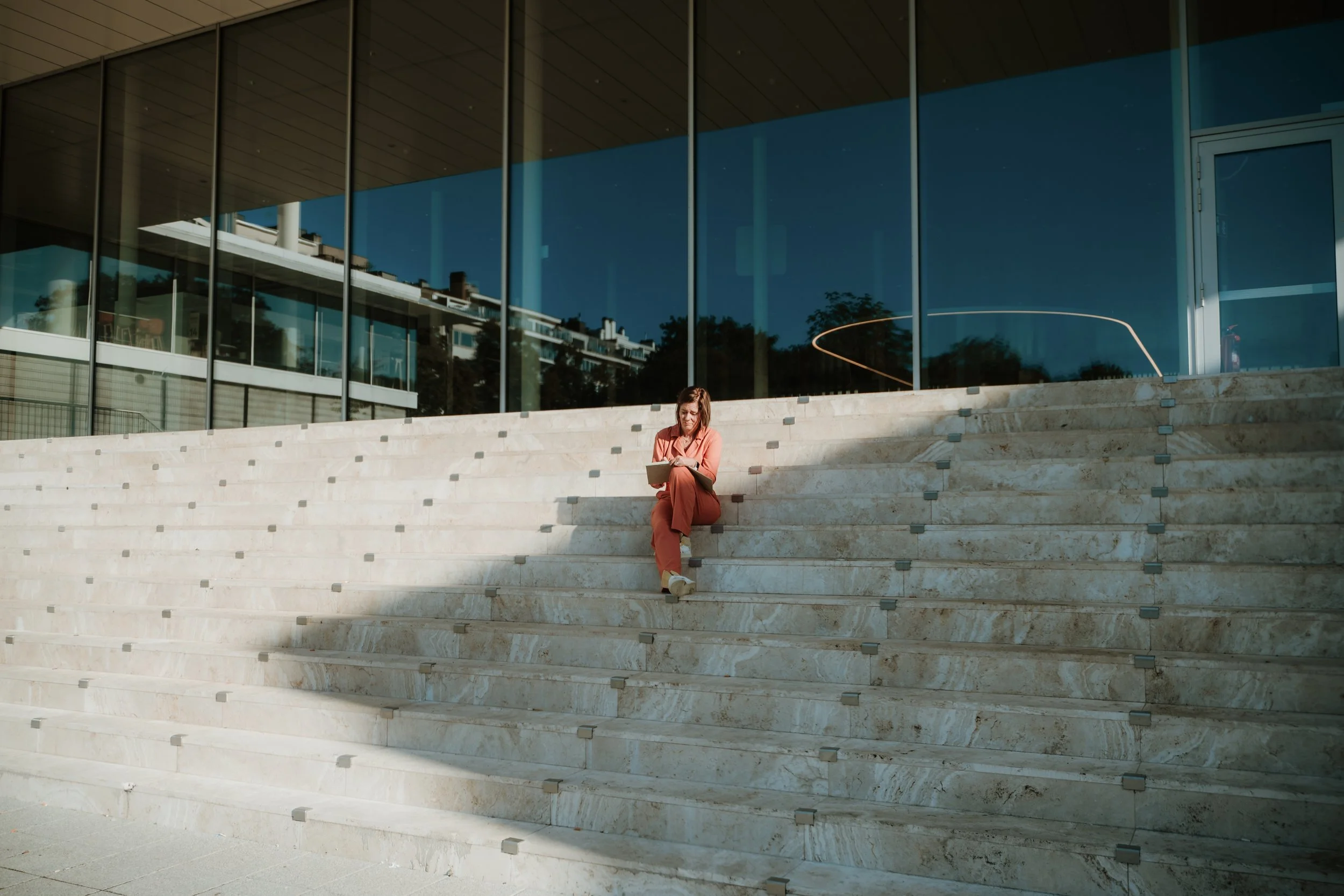 A woman sitting on wide marble steps outside a modern building with large glass windows, reading a book.