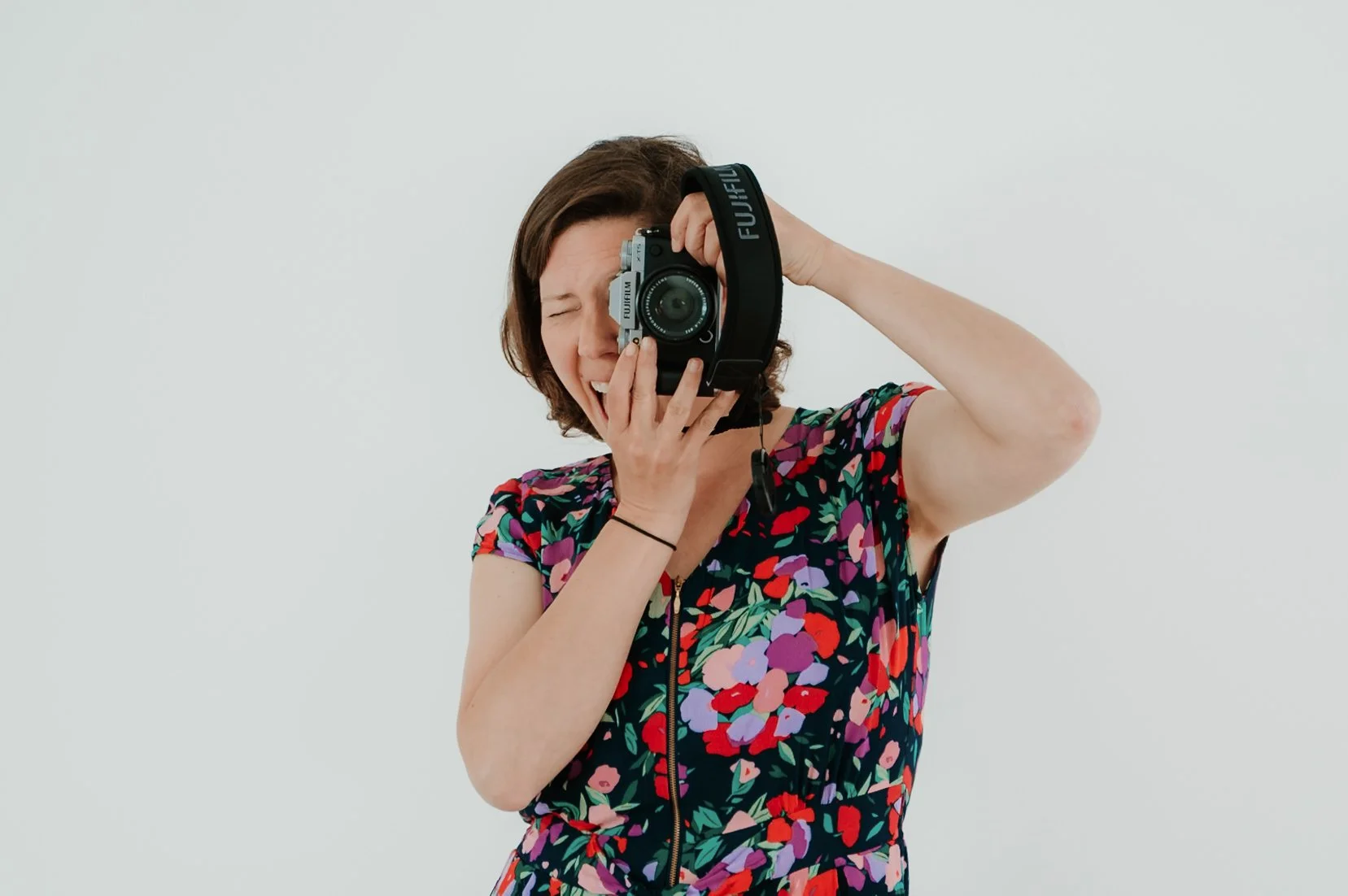 Woman with short brown hair in a floral dress taking a photo with a camera, looking through the viewfinder with one eye closed, against a plain white background.