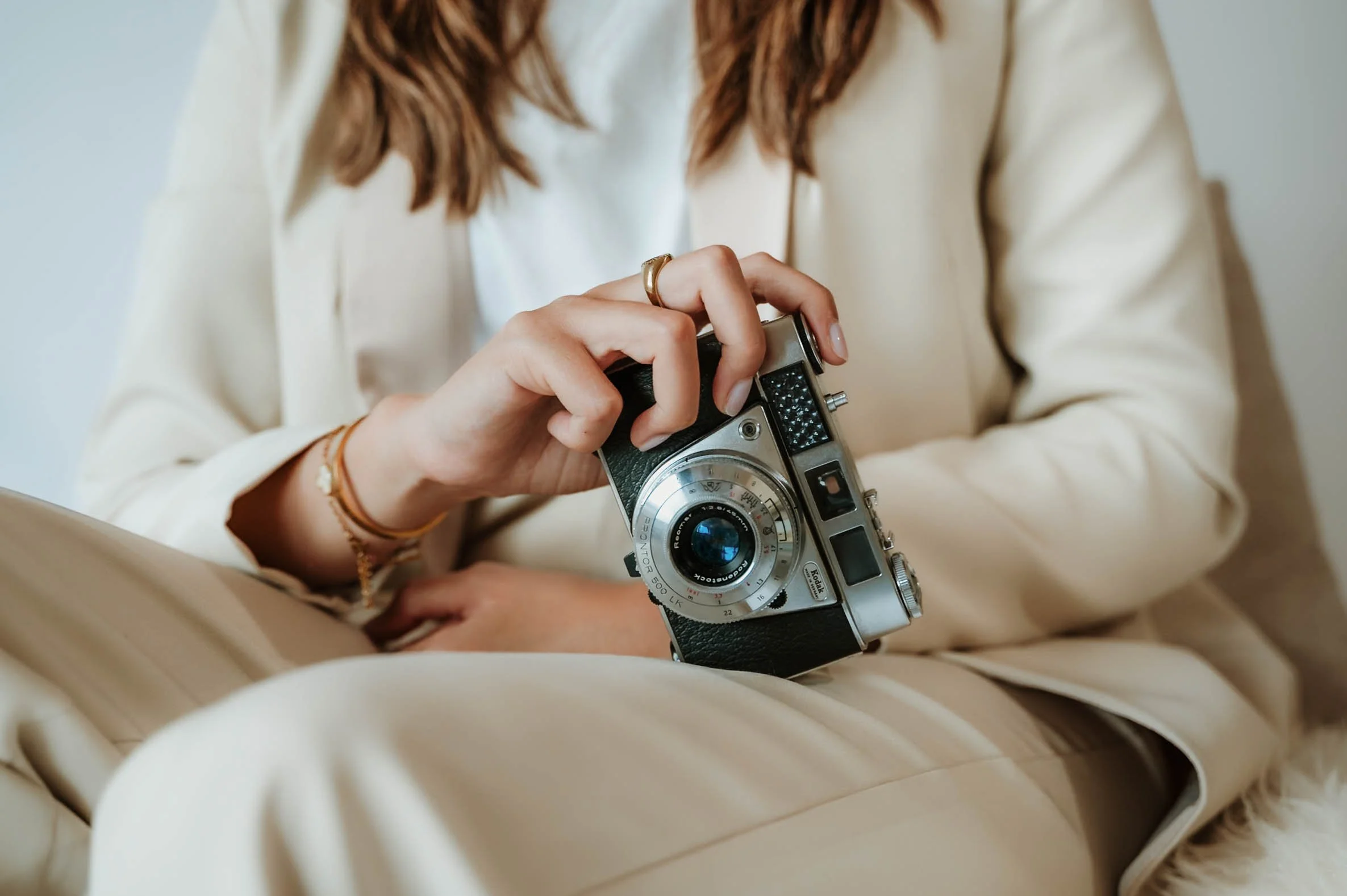 A woman is sitting on a beige couch, holding a vintage film camera in her hands. She is wearing a cream-colored blazer and has wavy, light brown hair. She is also wearing rings and a bracelet.