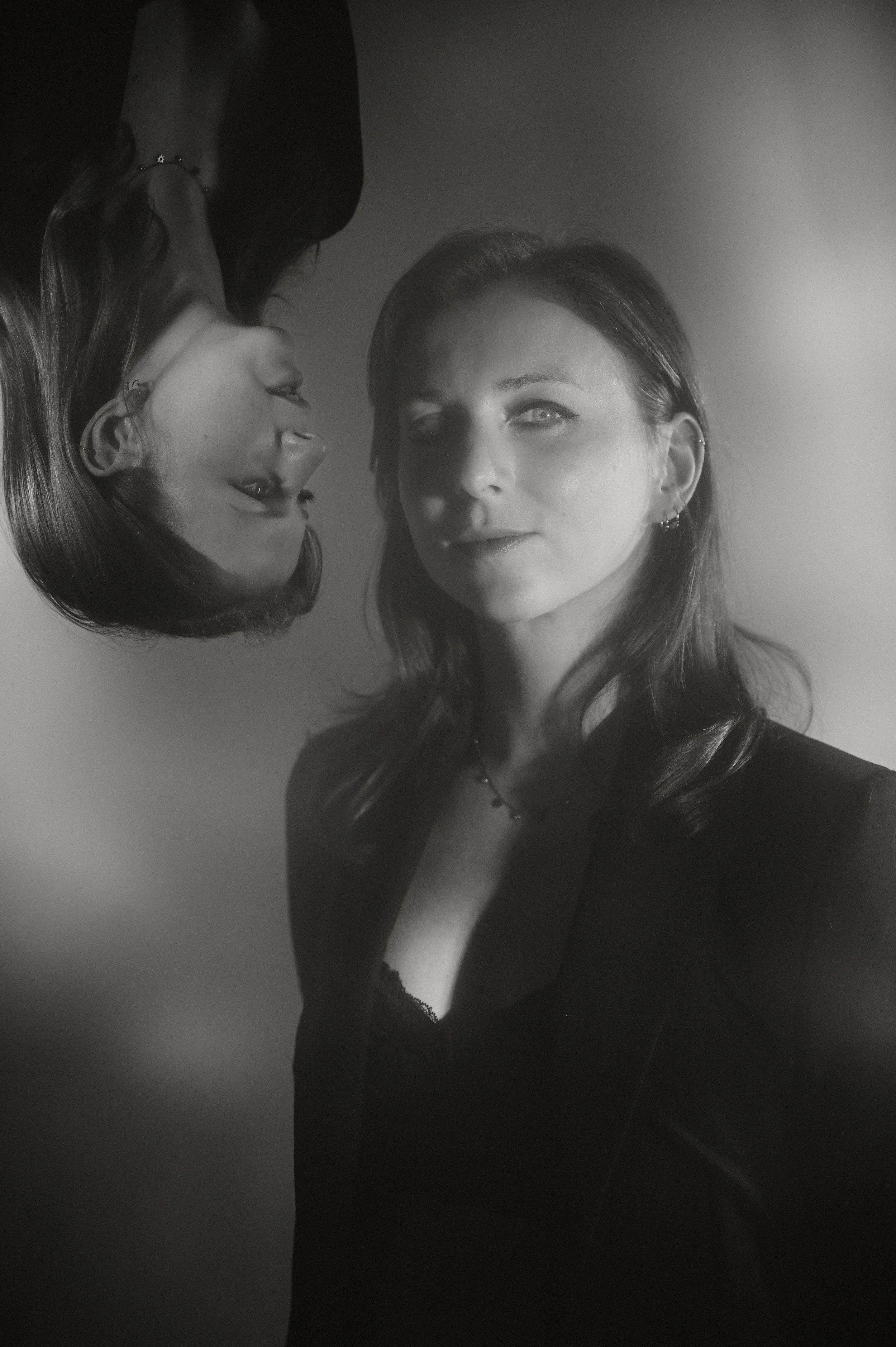 A black-and-white photograph featuring two women. One woman is seen in the foreground with long hair, earrings, and a necklace, wearing a low-cut top. The second woman, in the background, has shorter hair and is smiling, with her face reflected upsid