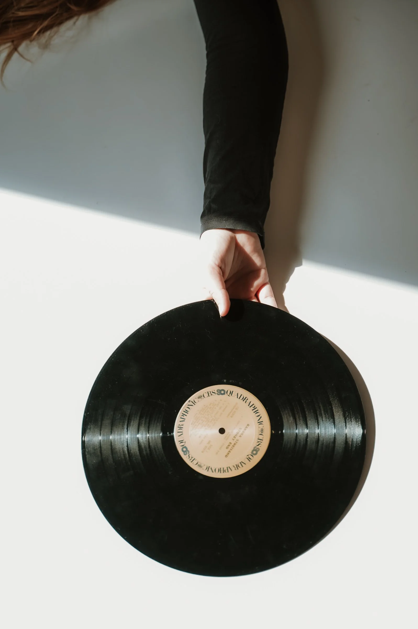 A person is holding a vinyl record by its edge with one hand while the record rests on a white surface. The person is wearing a black long-sleeve shirt. The vinyl record has a beige label with black text in the center.