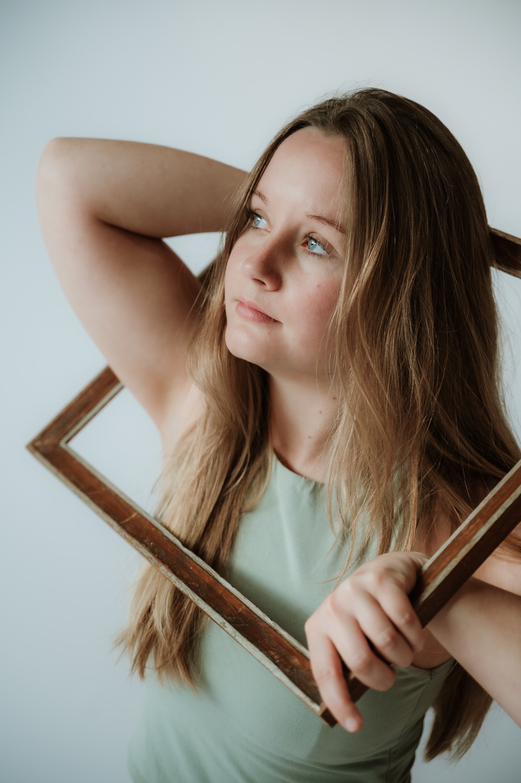 Young woman holding a vintage mirror, looking thoughtfully into the distance. The mirror reflects her face, showing her long, light brown hair and light-colored eyes. She is wearing a light green shirt and is against a plain white background.