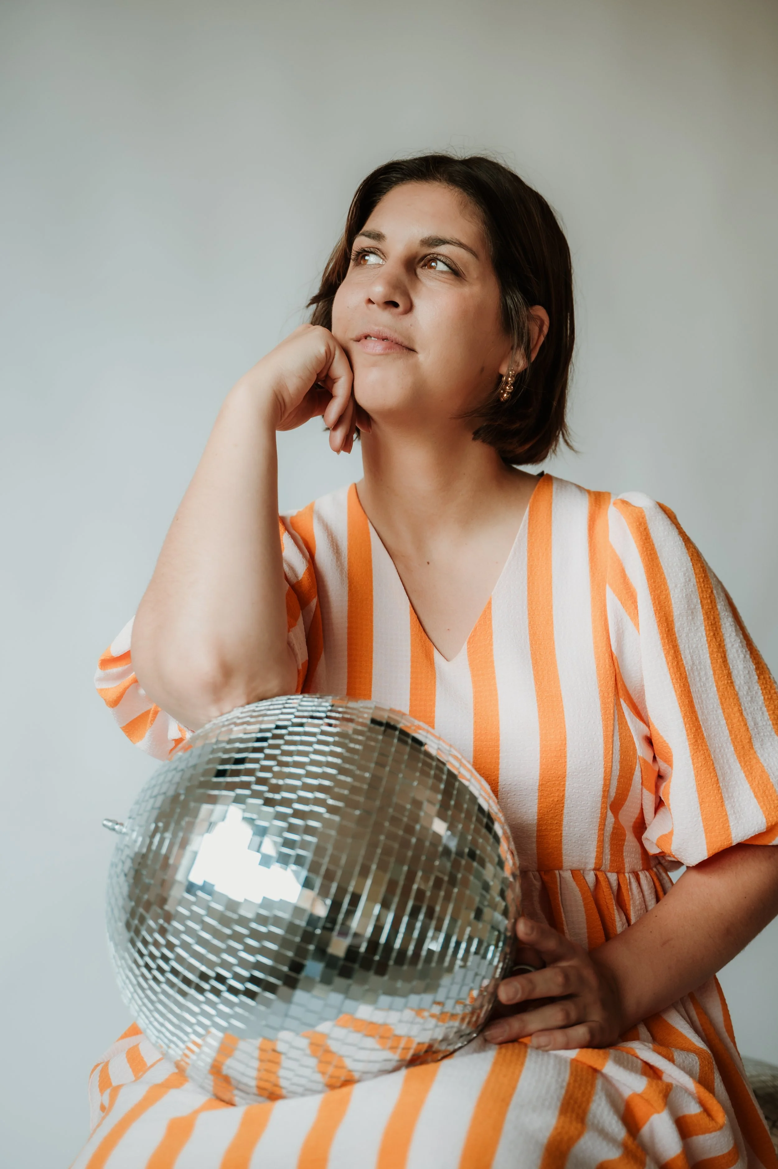 A woman wearing a striped orange and white dress is holding a small disco ball in front of her, one hand supporting the ball and the other resting on her chin, looking thoughtfully to the side with a slight smile.