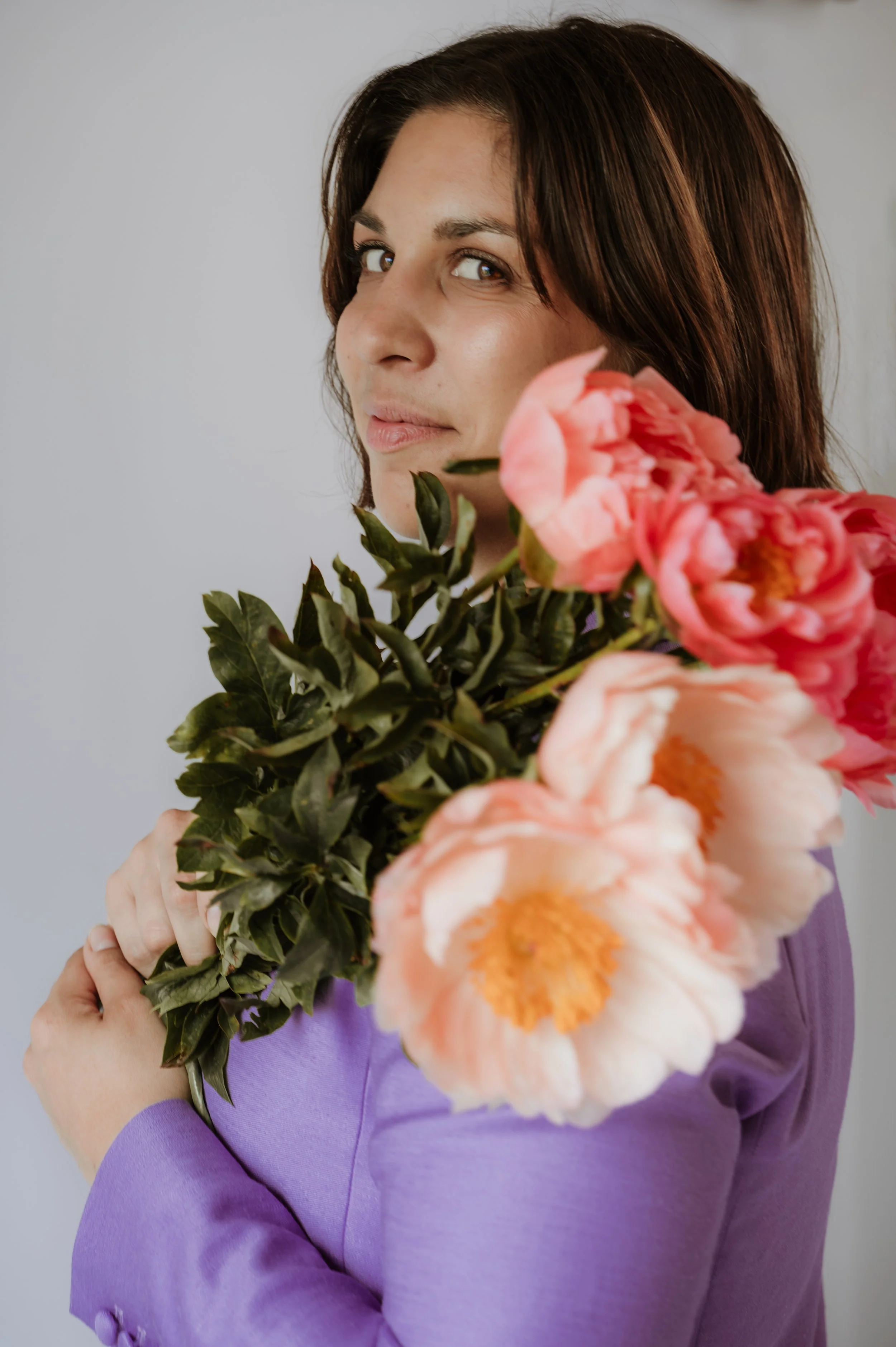 A woman with short brown hair and light skin, wearing a purple long-sleeve top, is holding a bouquet of pink and orange flowers close to her face. The background is plain and light-colored.