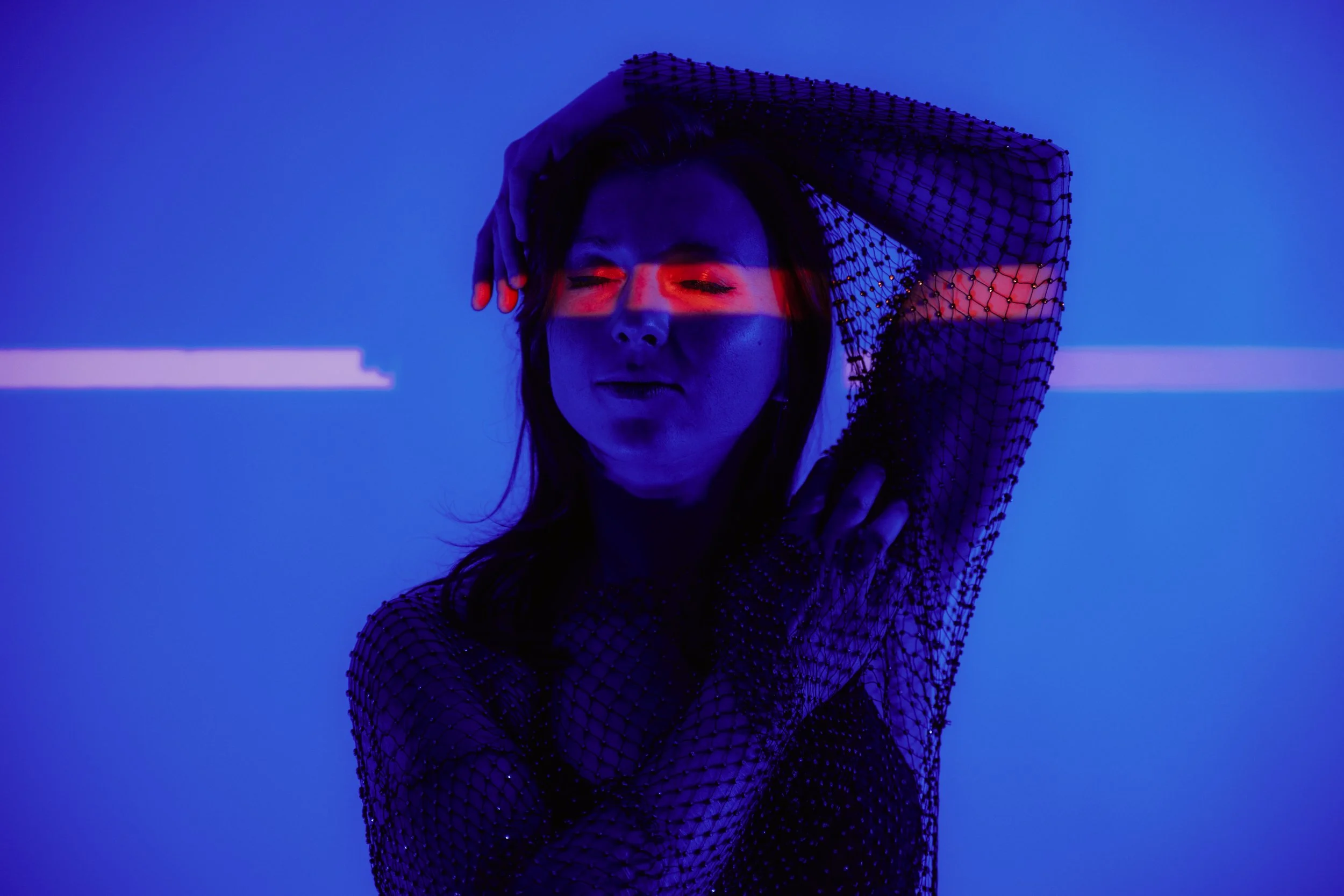 A woman with long hair posing with her hand above her head, wearing a black mesh top with small embellishments against a blue background with a red light across her eyes.