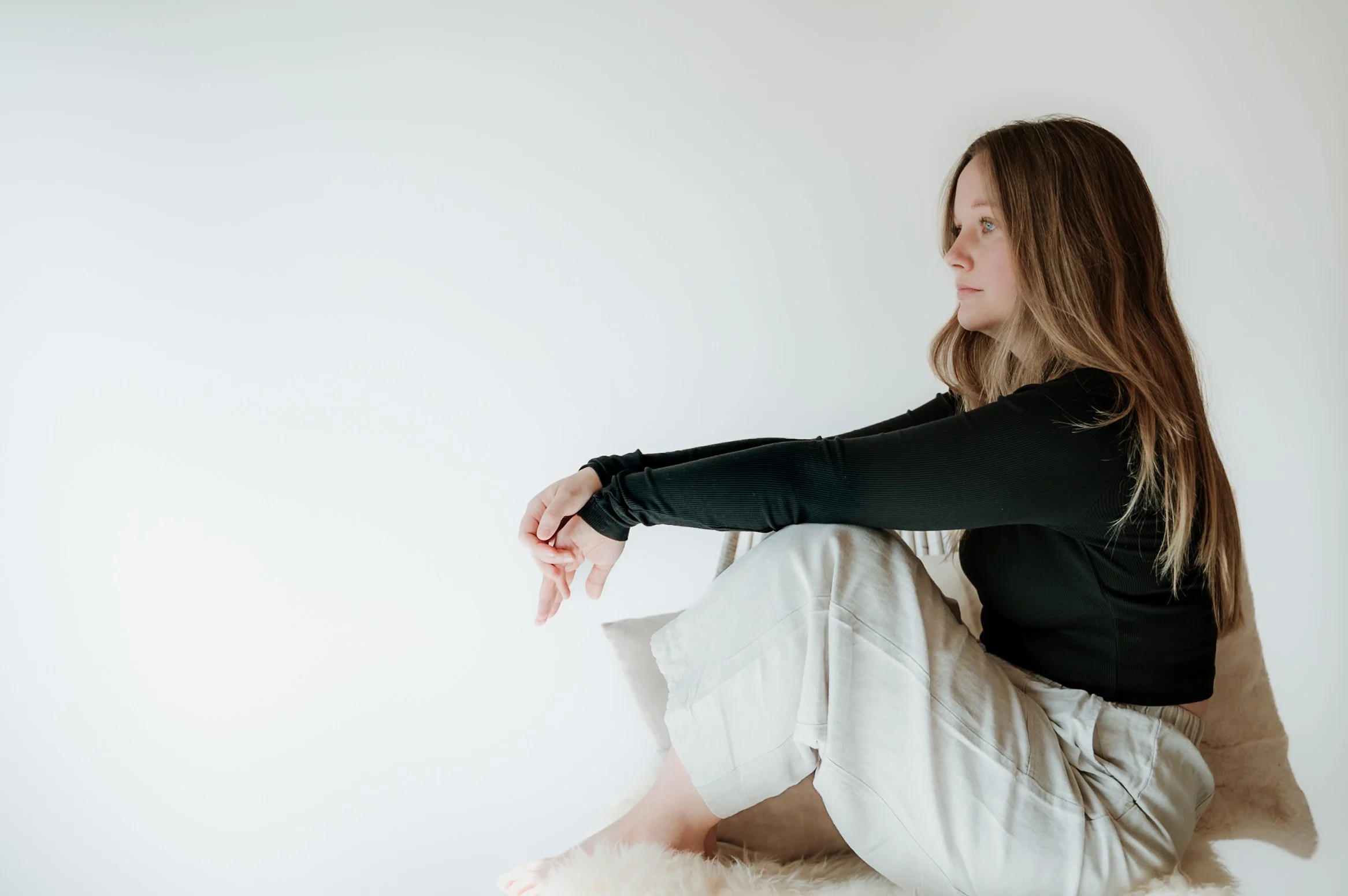 A woman sitting on the floor with her legs crossed, wearing a black long-sleeve top and light-colored pants, with light brown hair and blue eyes, looking to the right against a plain white background.