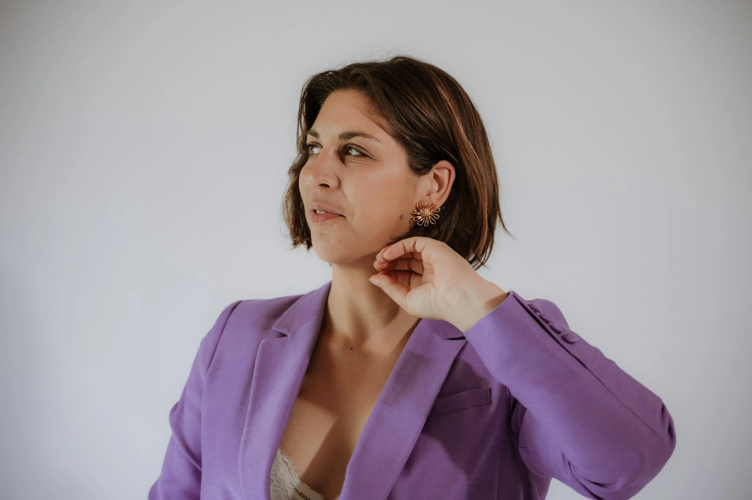 A woman with short brown hair wearing a purple blazer and floral earrings, posing against a plain light gray background.