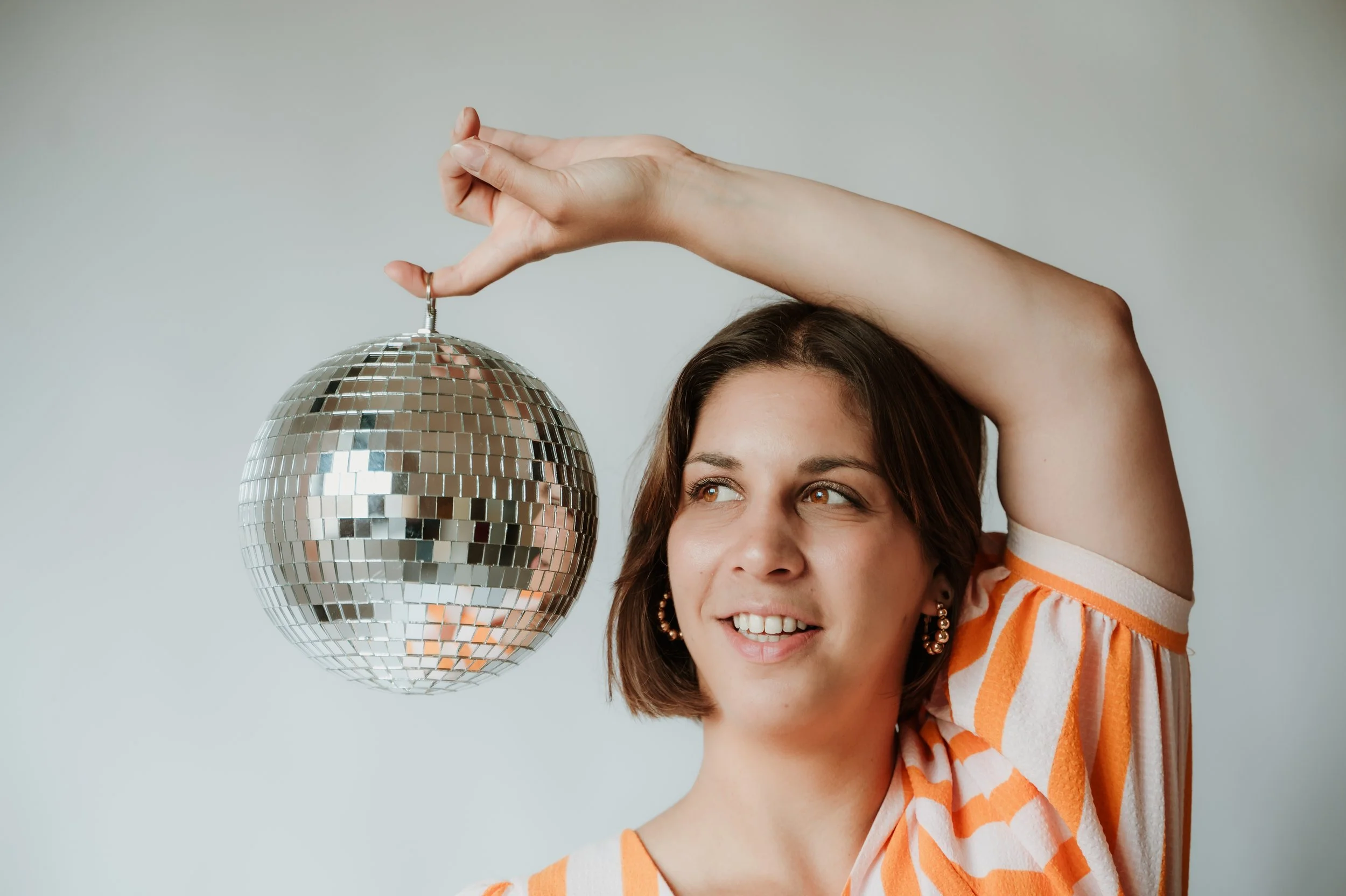 Woman with short brown hair and earrings smiling, holding a mirrored disco ball with her left hand. She is wearing a striped orange, white, and beige top, and is positioned against a plain light background.