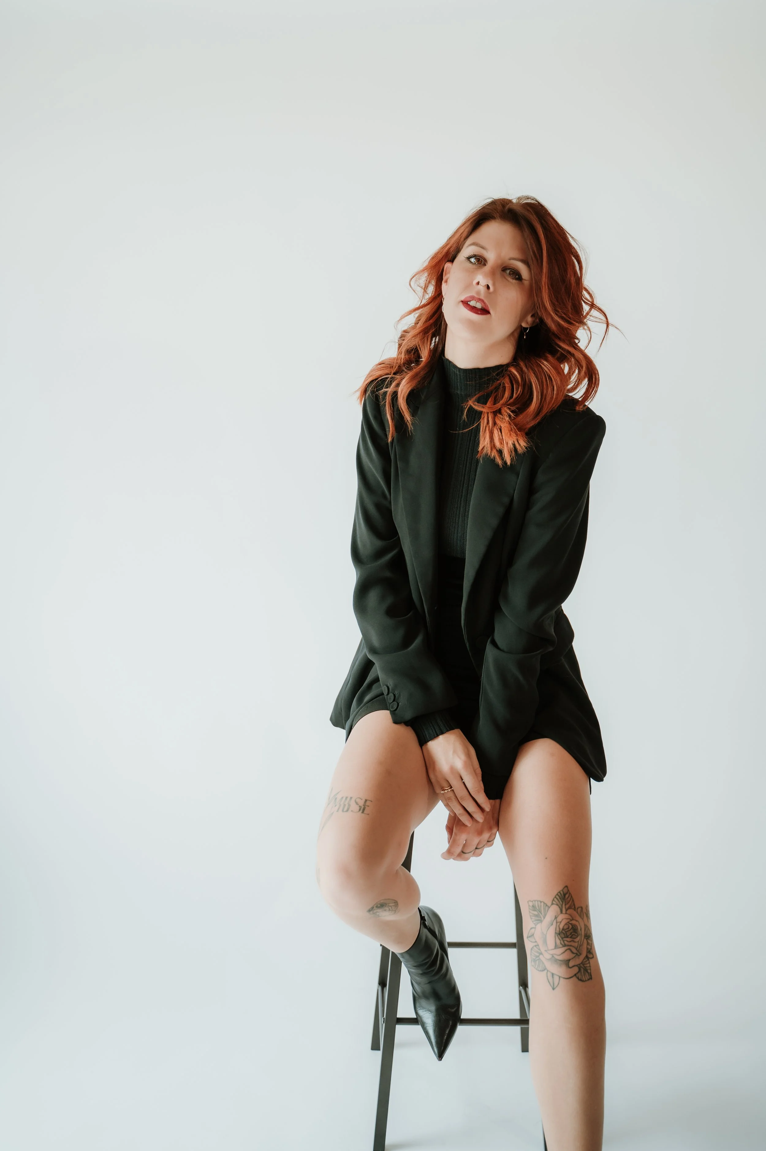 A woman with red hair and tattoos sitting on a stool against a plain white background, wearing a black blazer and boots, with her legs partially exposed.