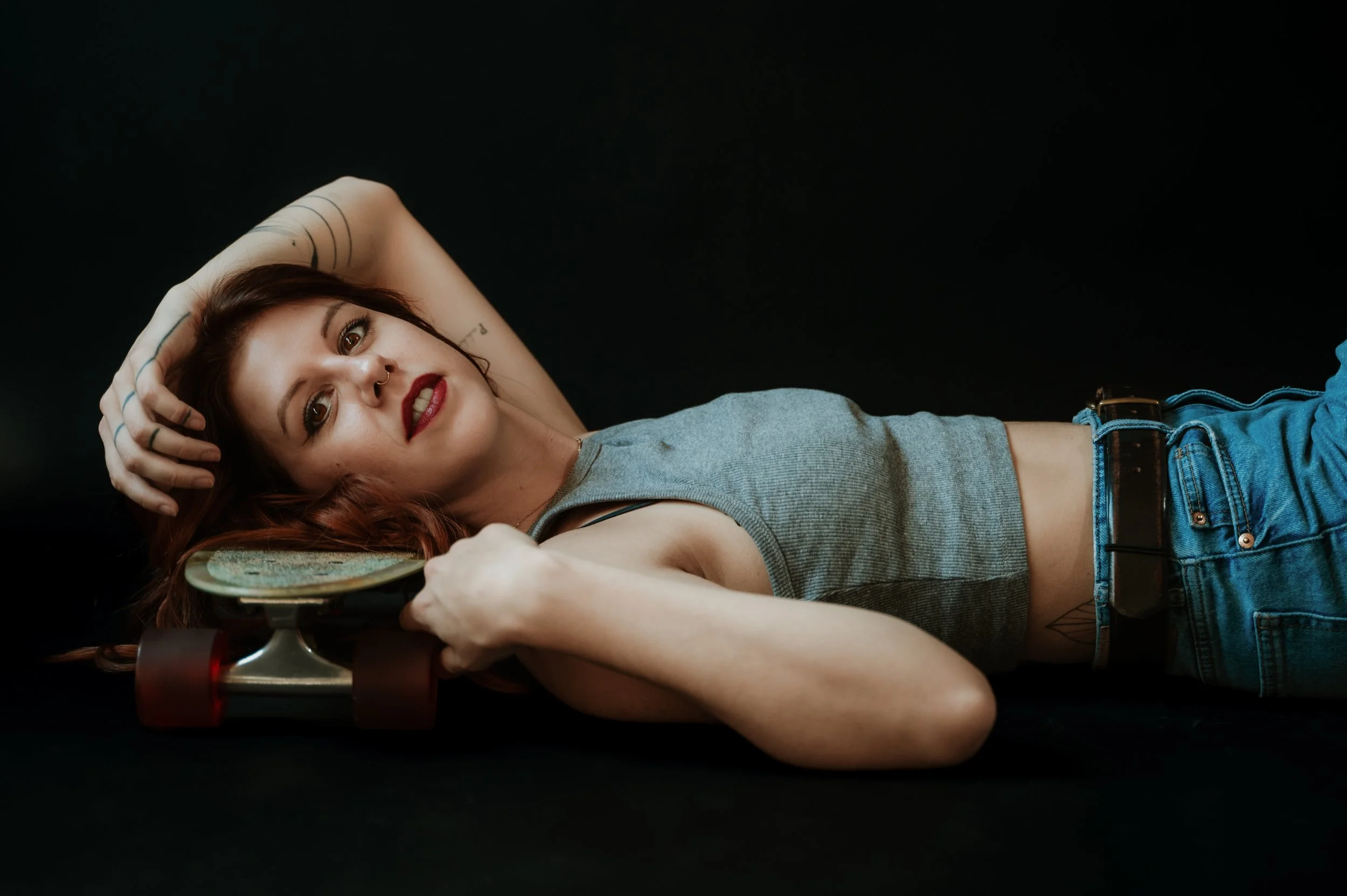 A woman with red hair, makeup, tattooed arms, and a septum piercing lying on a black background, holding a skateboard.