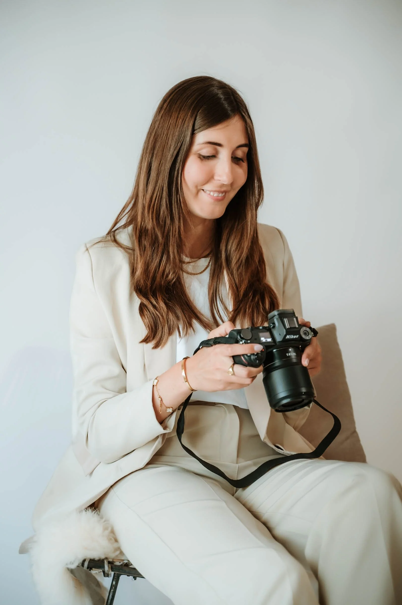 A young woman with long brown hair, dressed in a cream-colored blazer and pants, sitting on a chair with a white furry cushion, looking at and holding a black Nikon camera.