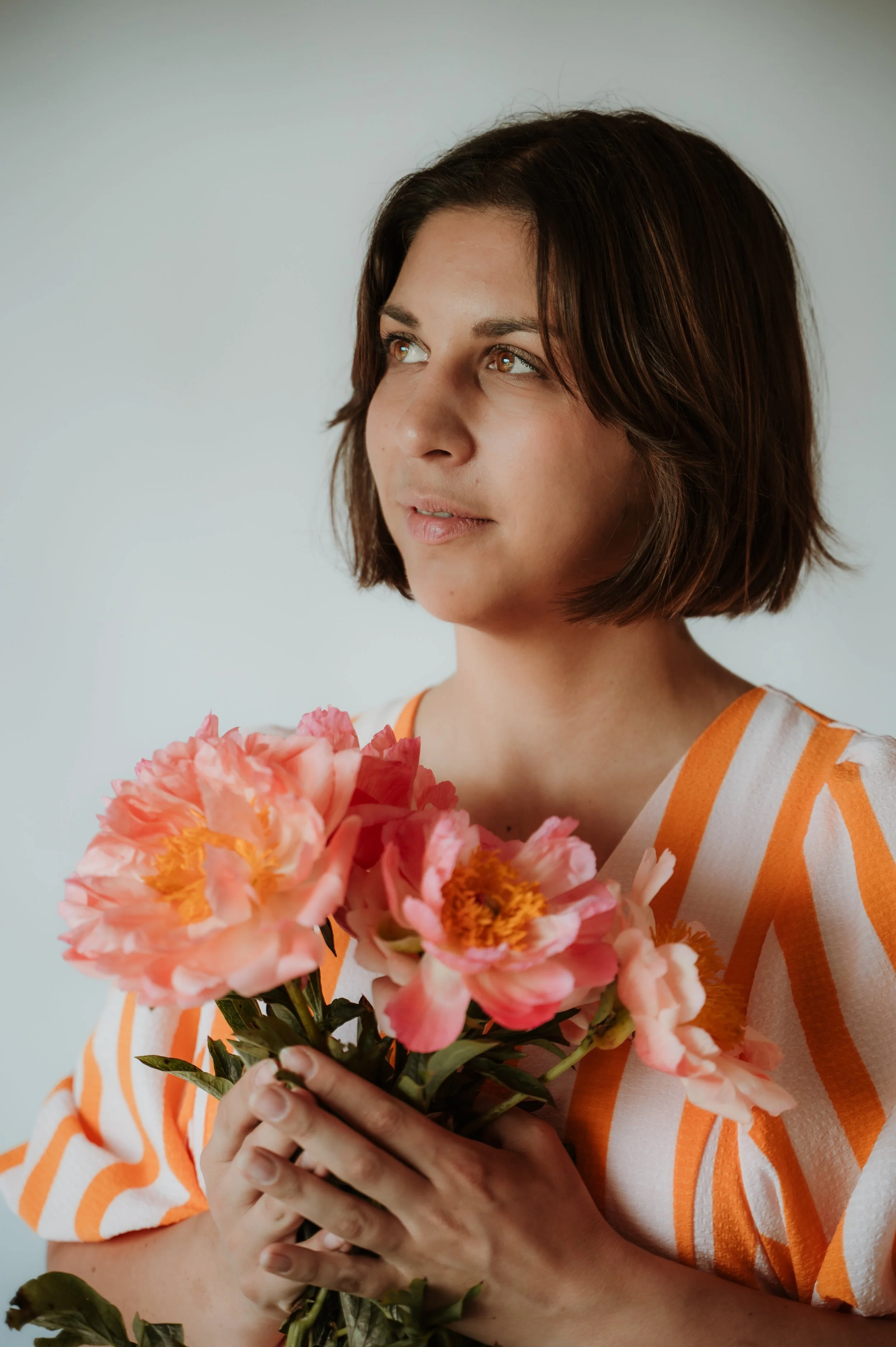A woman with short brown hair holding a bouquet of pink and orange flowers, wearing a white and orange striped shirt, looking to the side.