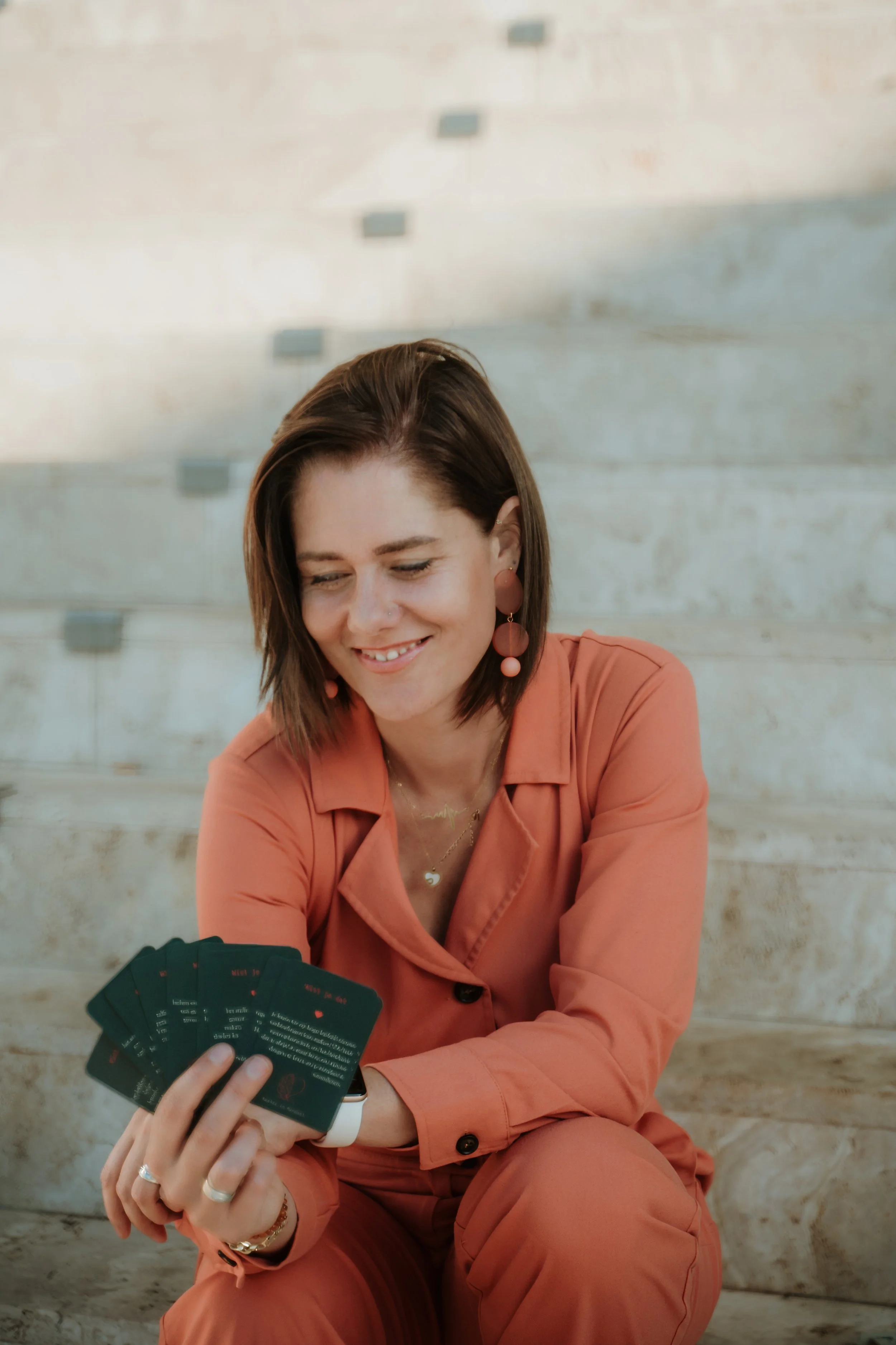 A woman with brown hair, dressed in a coral-colored suit, sitting on stone stairs, holding and showing a set of black cards with red and white text, smiling and looking down at the cards.