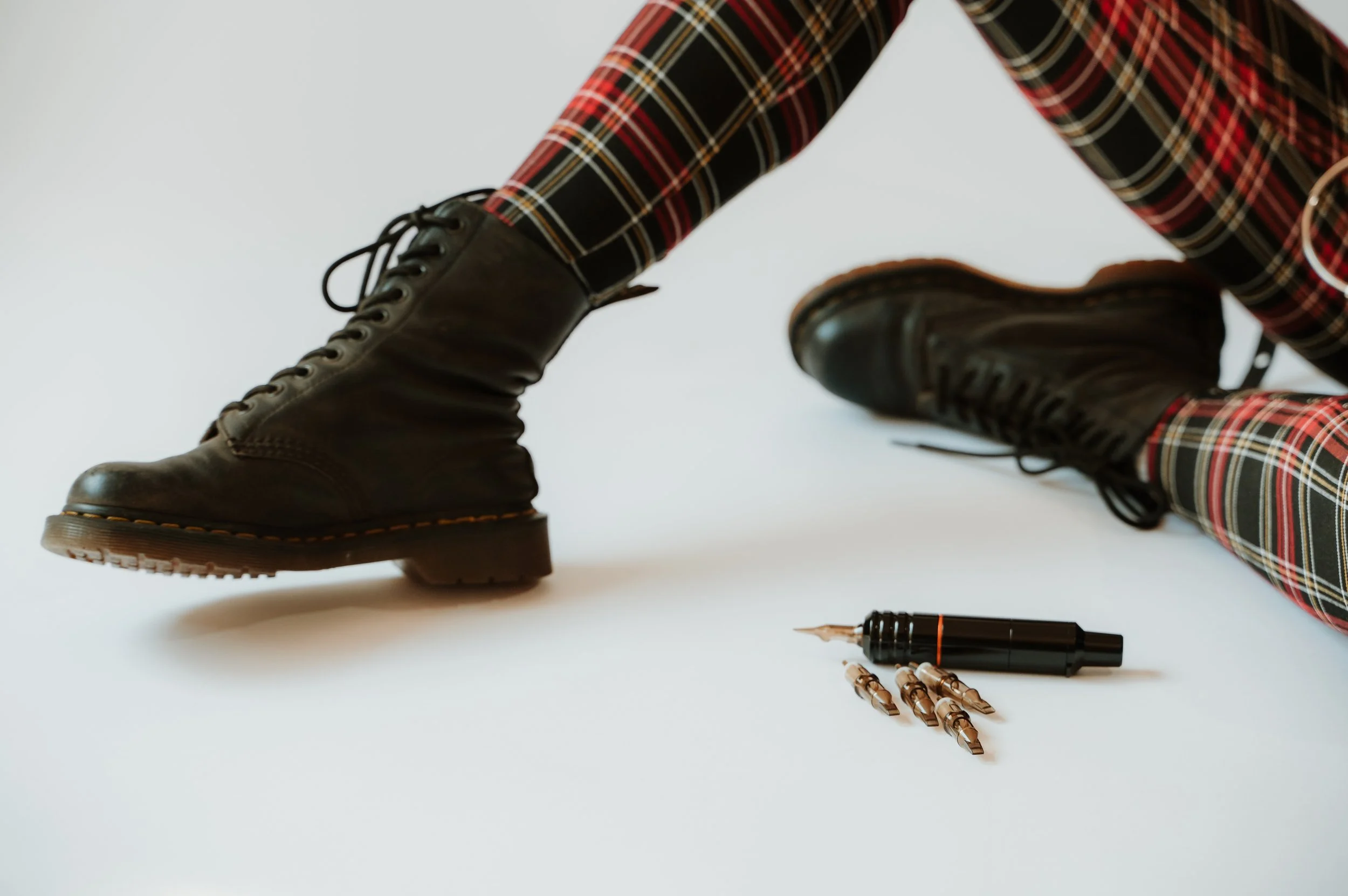 Close-up of black combat boots, plaid pants, black pen, and gold pens on white surface.