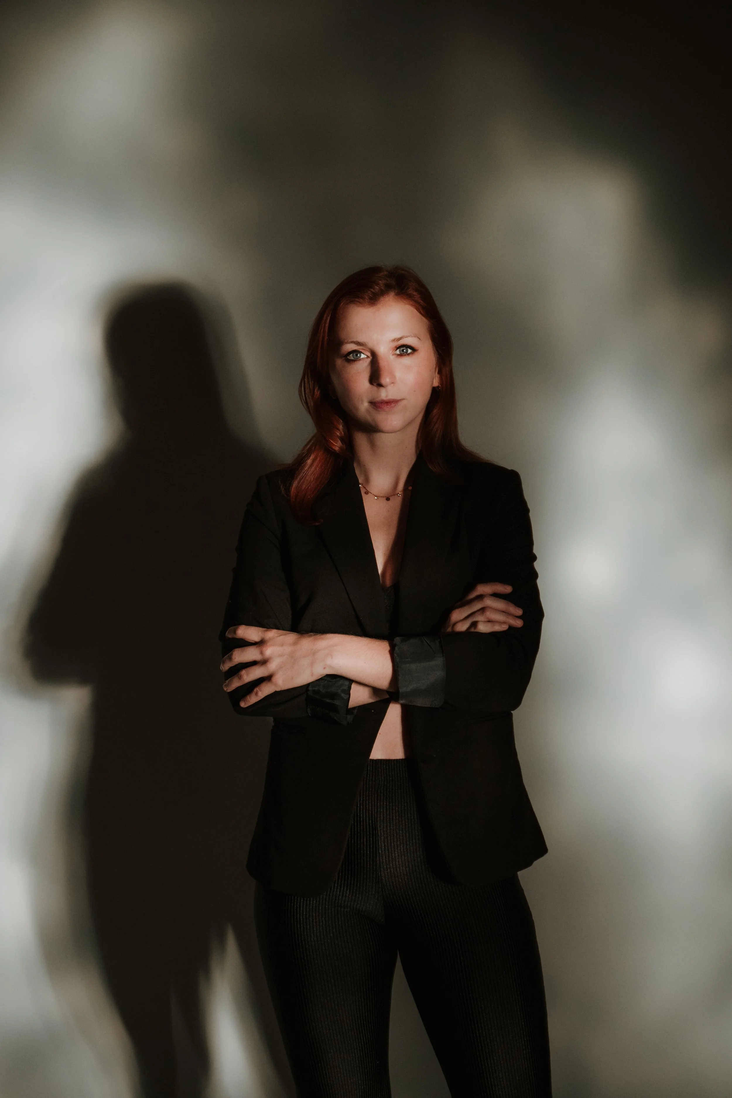A woman with red hair standing with arms crossed, wearing a black blazer and black pants, in a dimly lit room with shadows cast on the wall behind her.