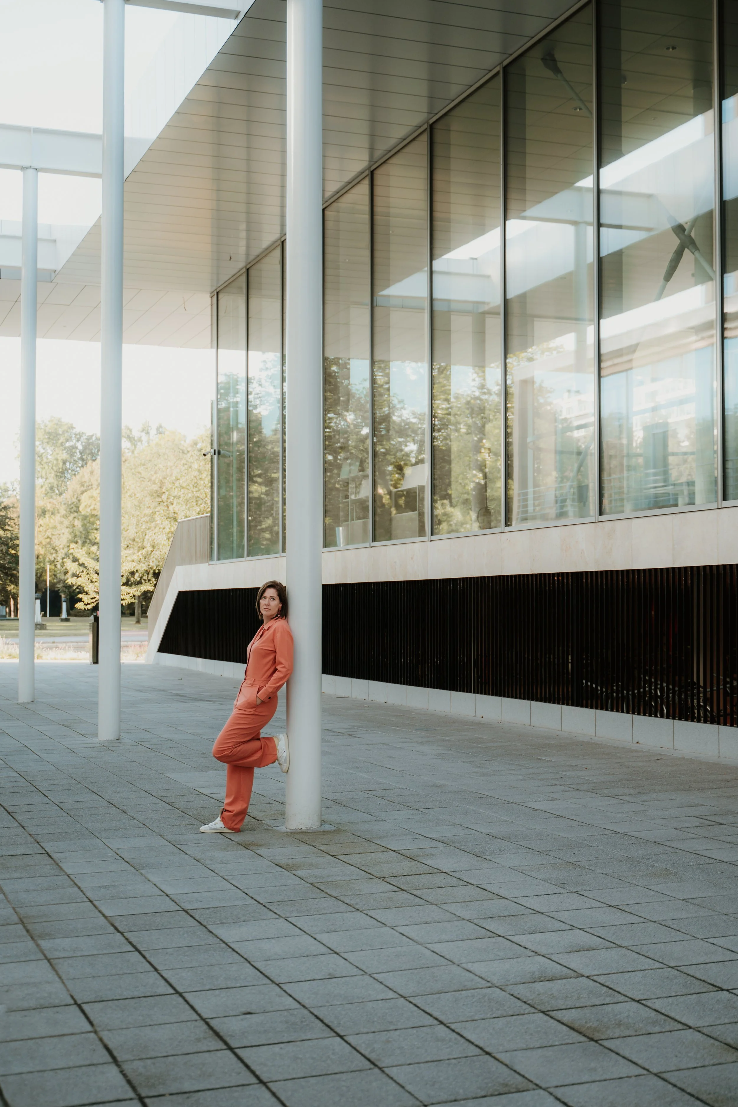 A woman in an orange jumpsuit leaning against a white pole outside modern glass building with reflective windows.
