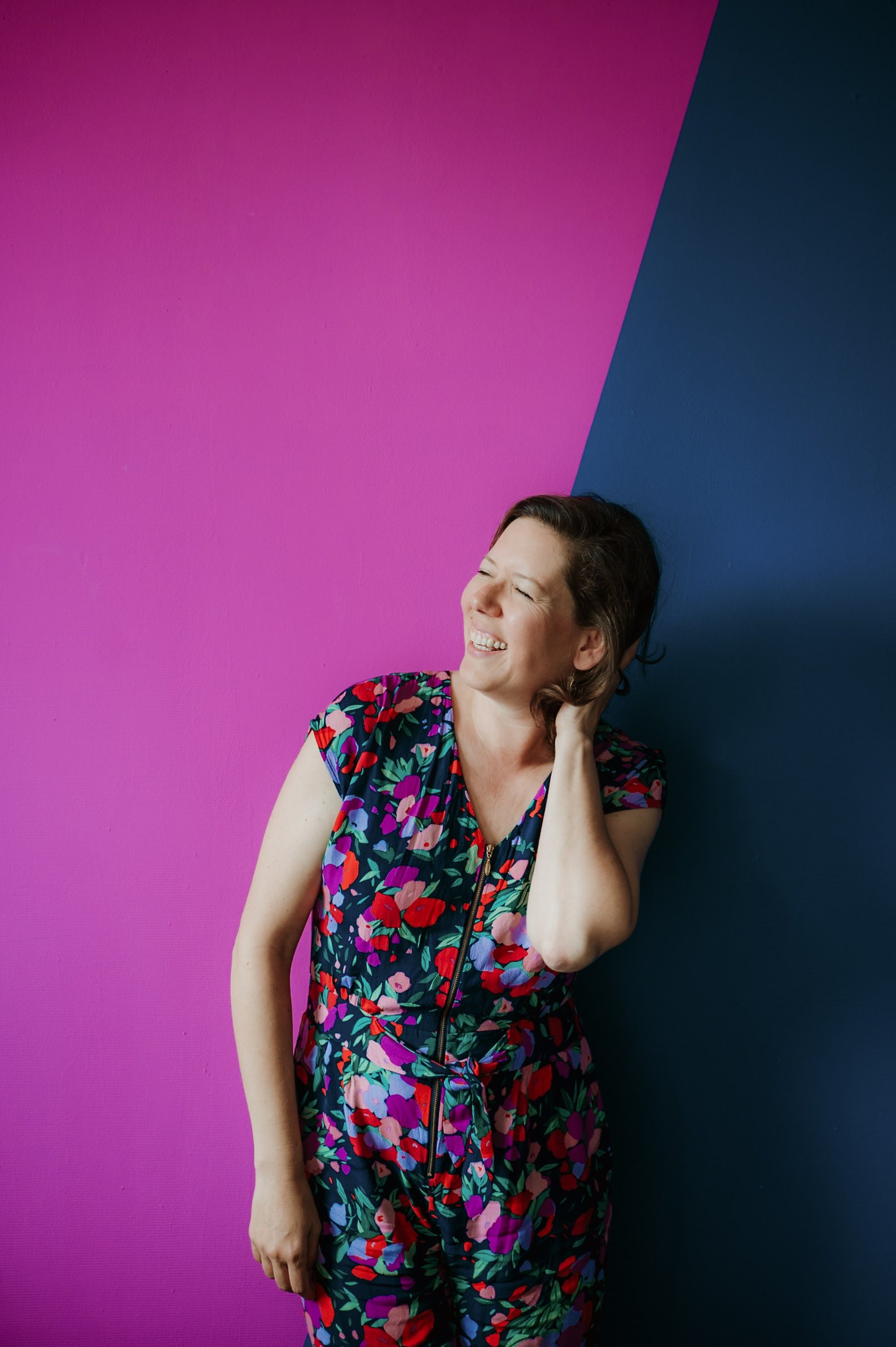 A woman with short hair is smiling and leaning her head against a wall that is painted pink and dark blue, wearing a colorful floral dress with a zipper.