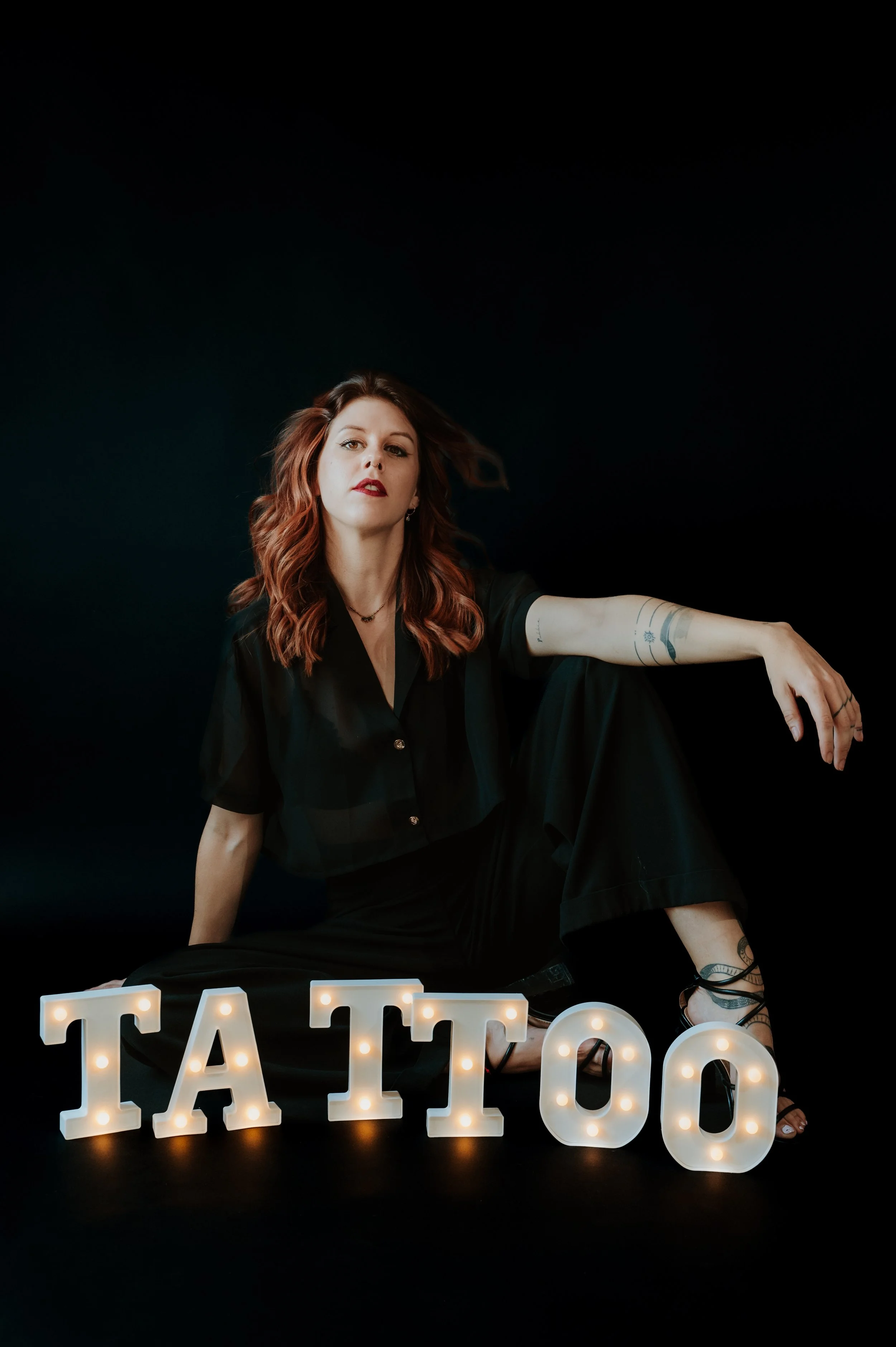 A woman with wavy red hair and tattoos on her arm, wearing a black outfit, sitting against a black background with illuminated marquee letters spelling 'TATTOOS' in front of her.