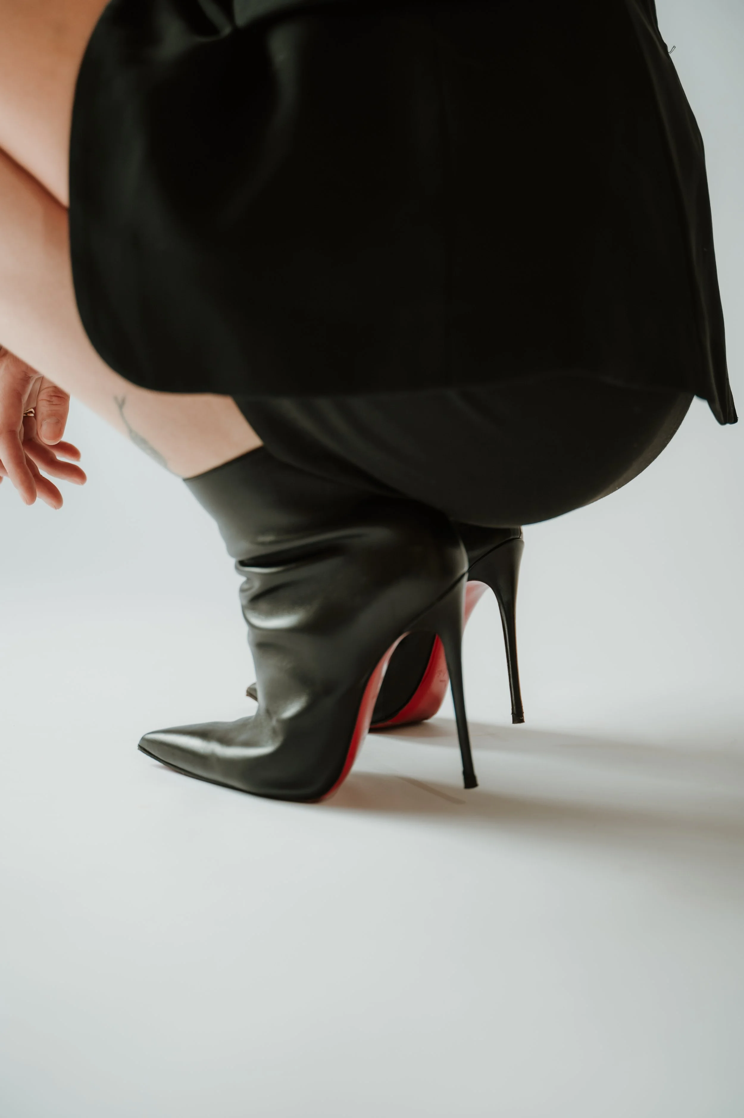 Close-up of a person's legs wearing black high-heeled boots, with a black handbag resting on their lap, taken from a low angle against a plain white background.