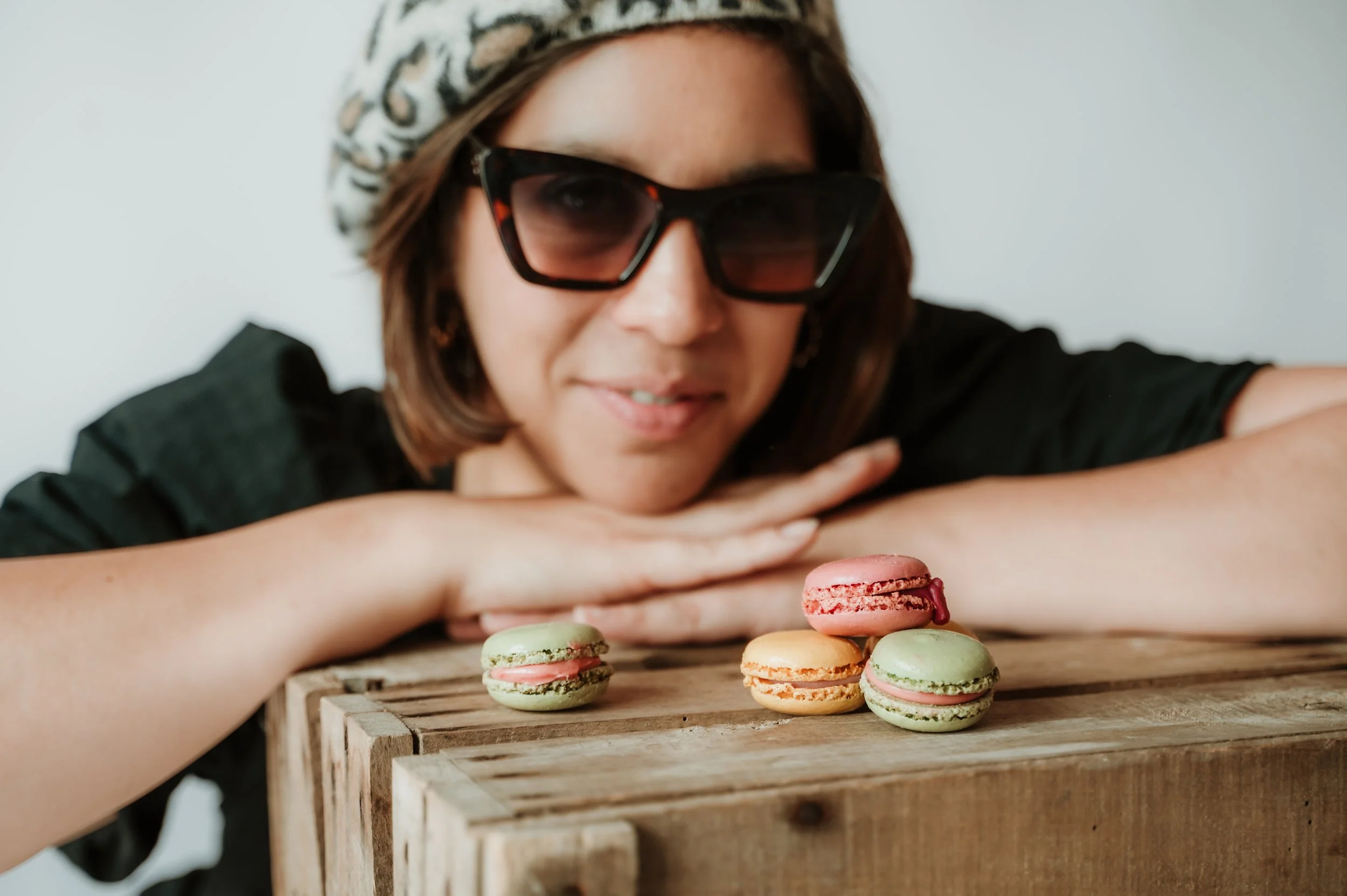 A woman wearing sunglasses and a headscarf resting her head on her arms in front of a wooden box filled with colorful macarons.