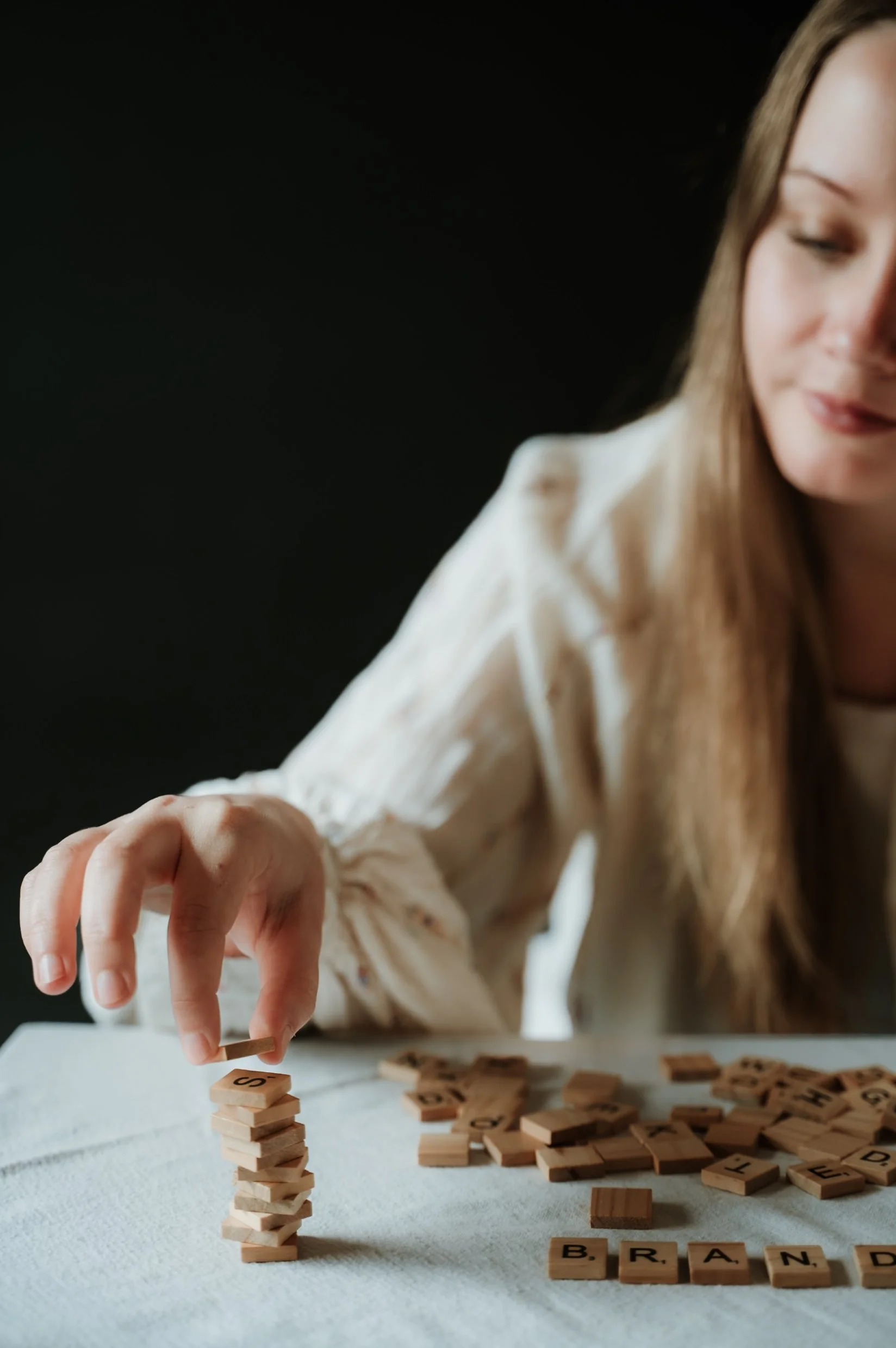 A young woman with long brown hair wearing a white shirt playing Scrabble, stacking tiles to build a tower, with the word 'BRAND' spelled out on tiles on the table.