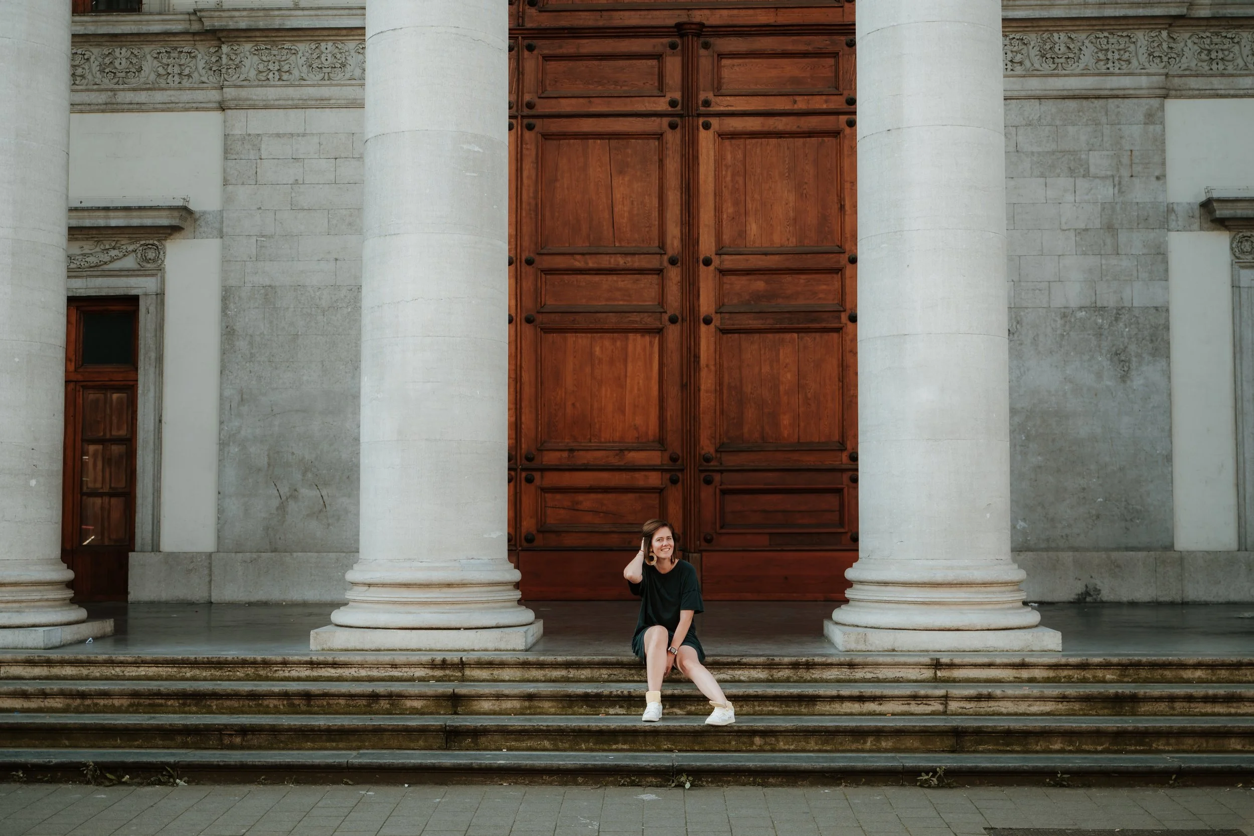 A woman with long hair, wearing a black top and white shoes, sitting on steps in front of a large building with tall white columns and a wooden door.