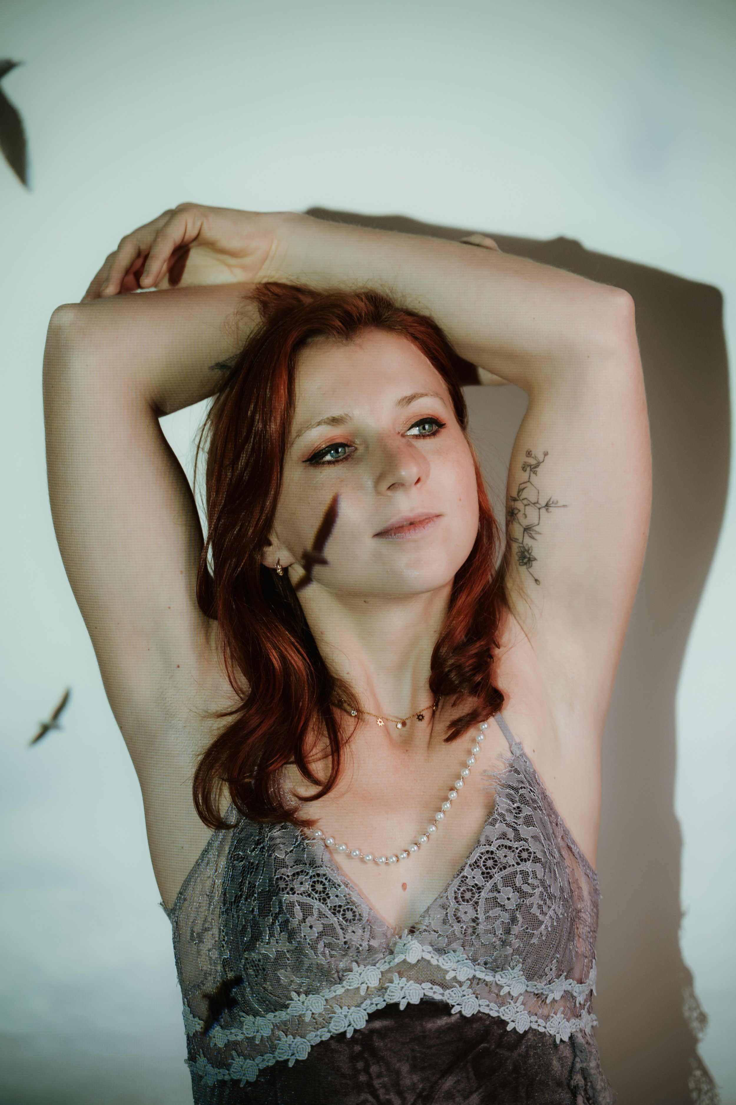 A woman with red hair and blue eyes, wearing a grey lace top, a pearl necklace, and a floral tattoo on her arm, poses with her arms raised above her head in front of a light-colored background.