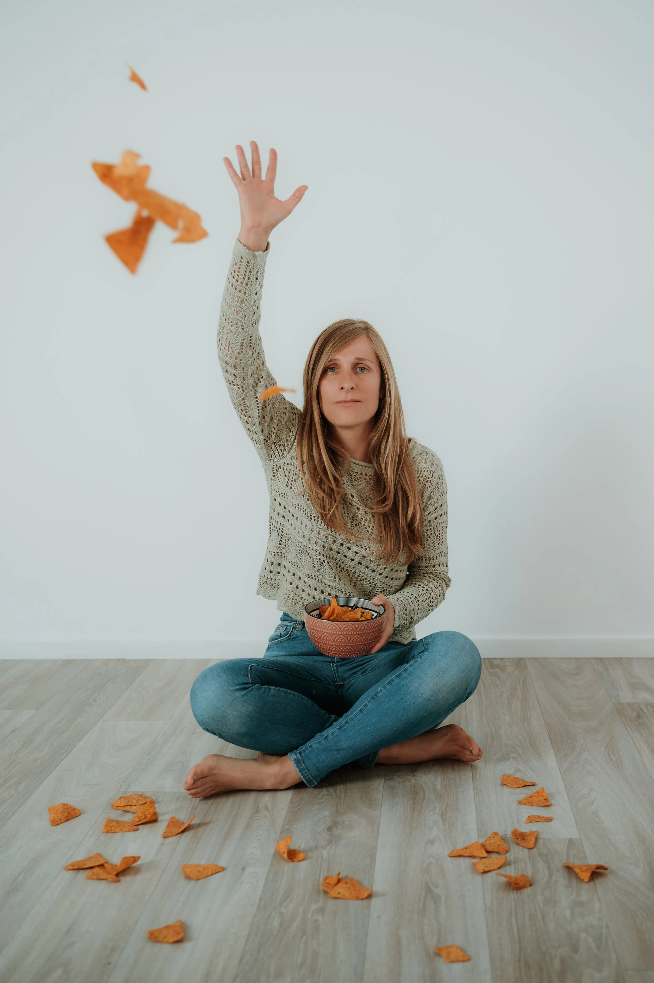 A woman sitting cross-legged on a wooden floor, holding a bowl of nachos, with some nacho chips scattered around her. She is reaching up to catch a falling nacho chip. The background is a plain, light-colored wall.