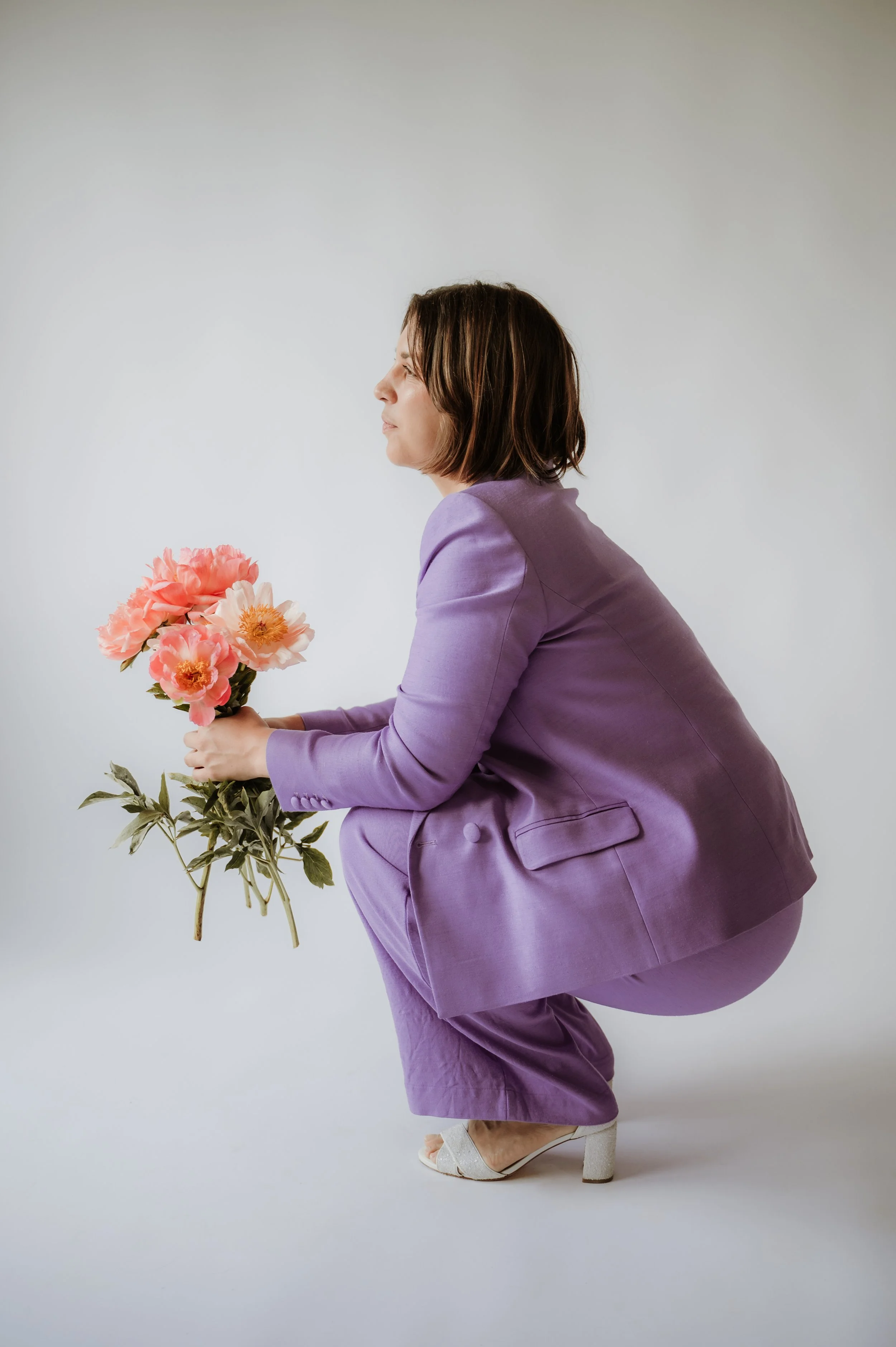 Woman in purple pants and blazer holding a bouquet of pink flowers, standing against a plain white background.