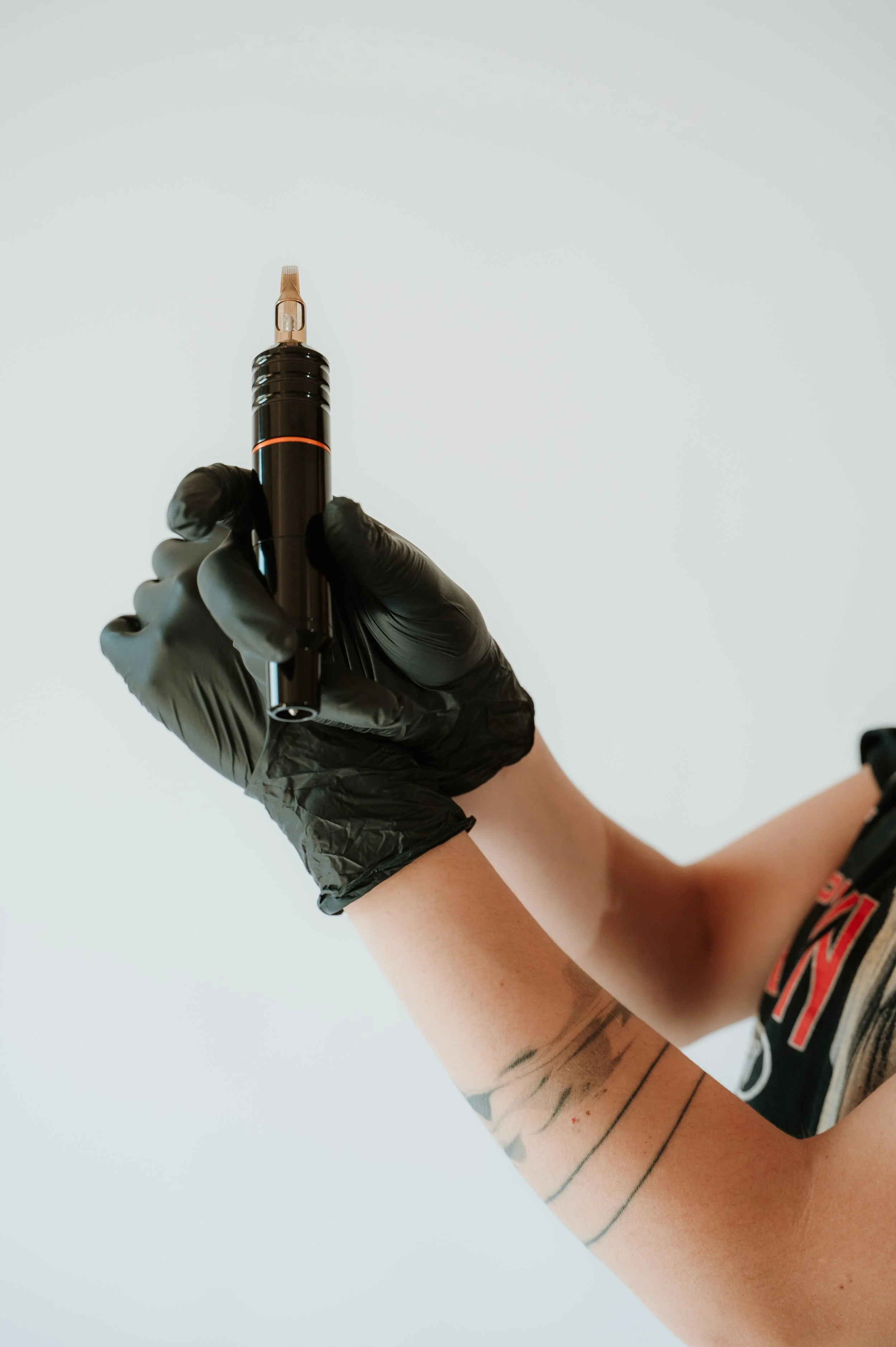 Person with tattooed arm wearing a black glove holding a pen or tattoo machine against a plain light background.