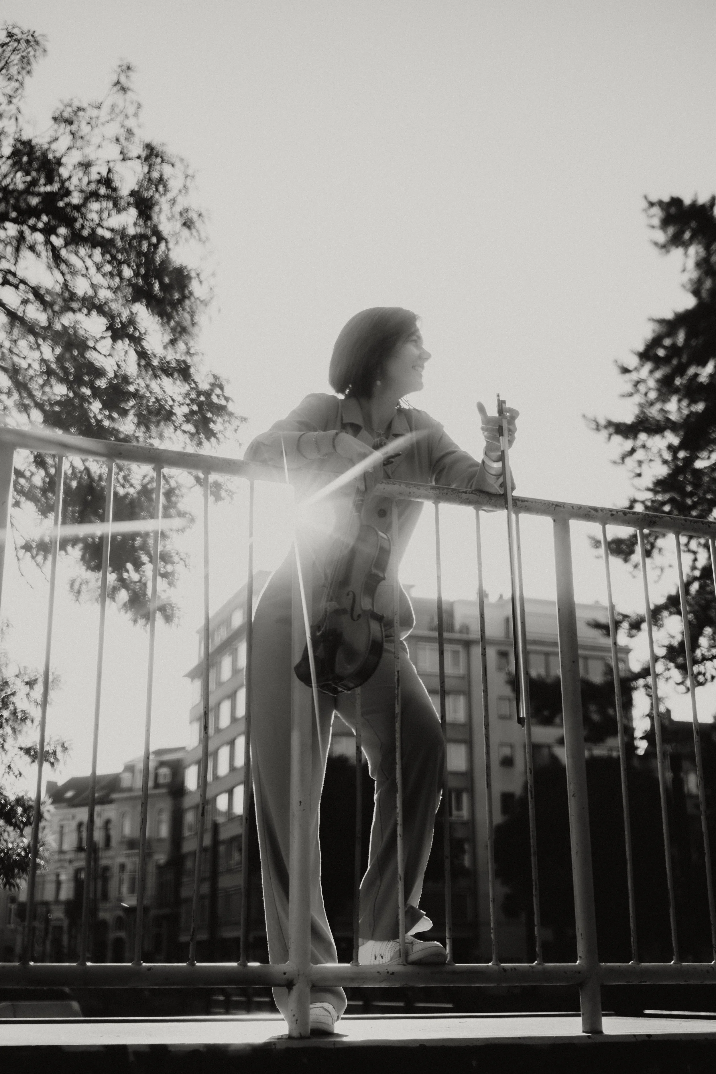 A woman smiling and holding a violin case, standing behind a metal railing outdoors with several trees and a building in the background.